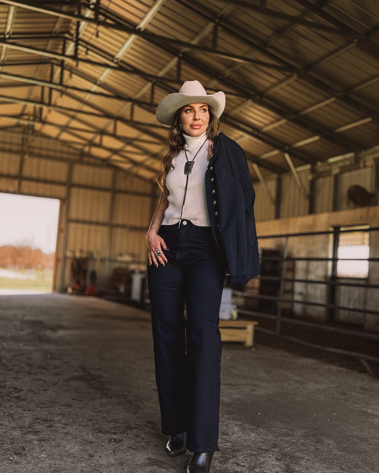Woman in a white cowboy hat and western boho outfit walking in a barn, NFR style fashion.