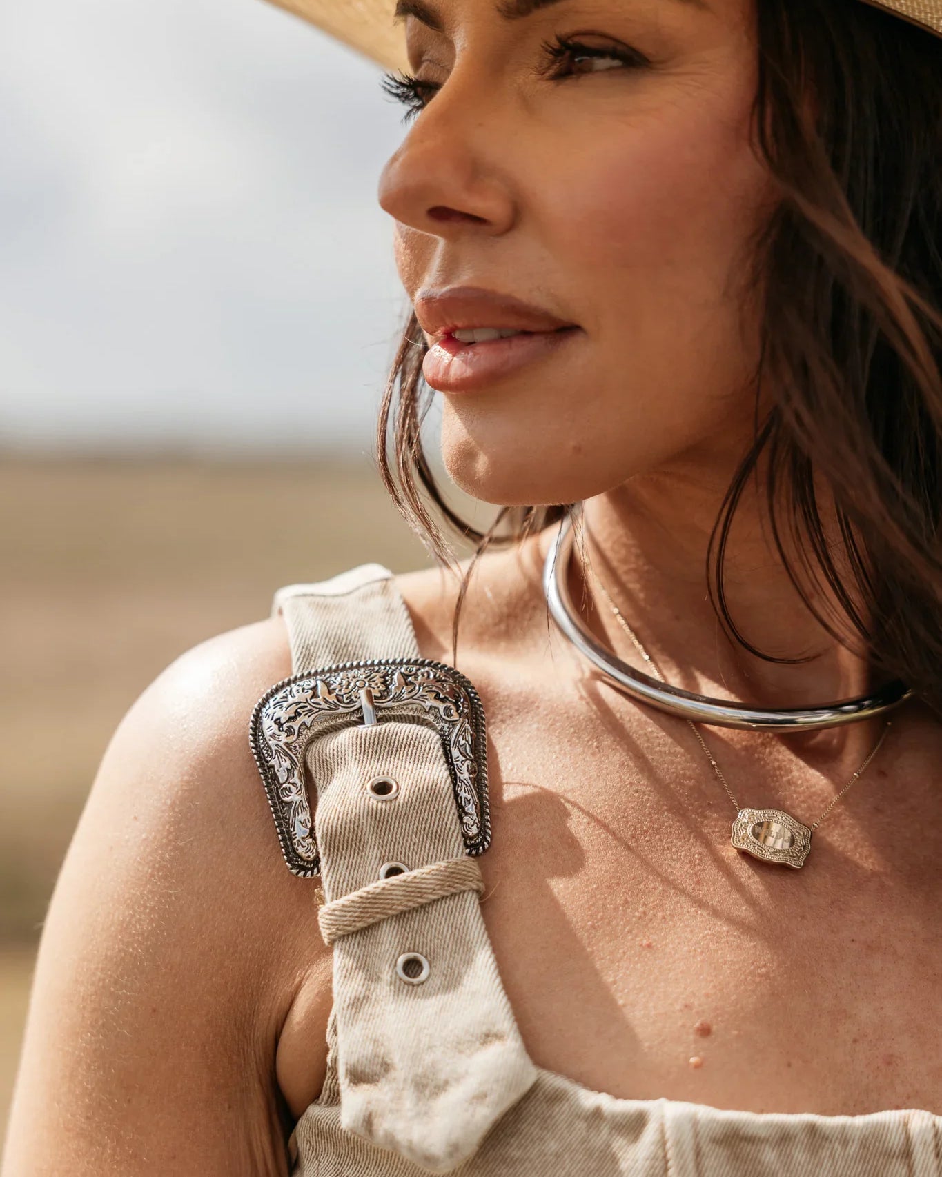 Woman wearing a beige hat and overalls with a necklace, standing outdoors.