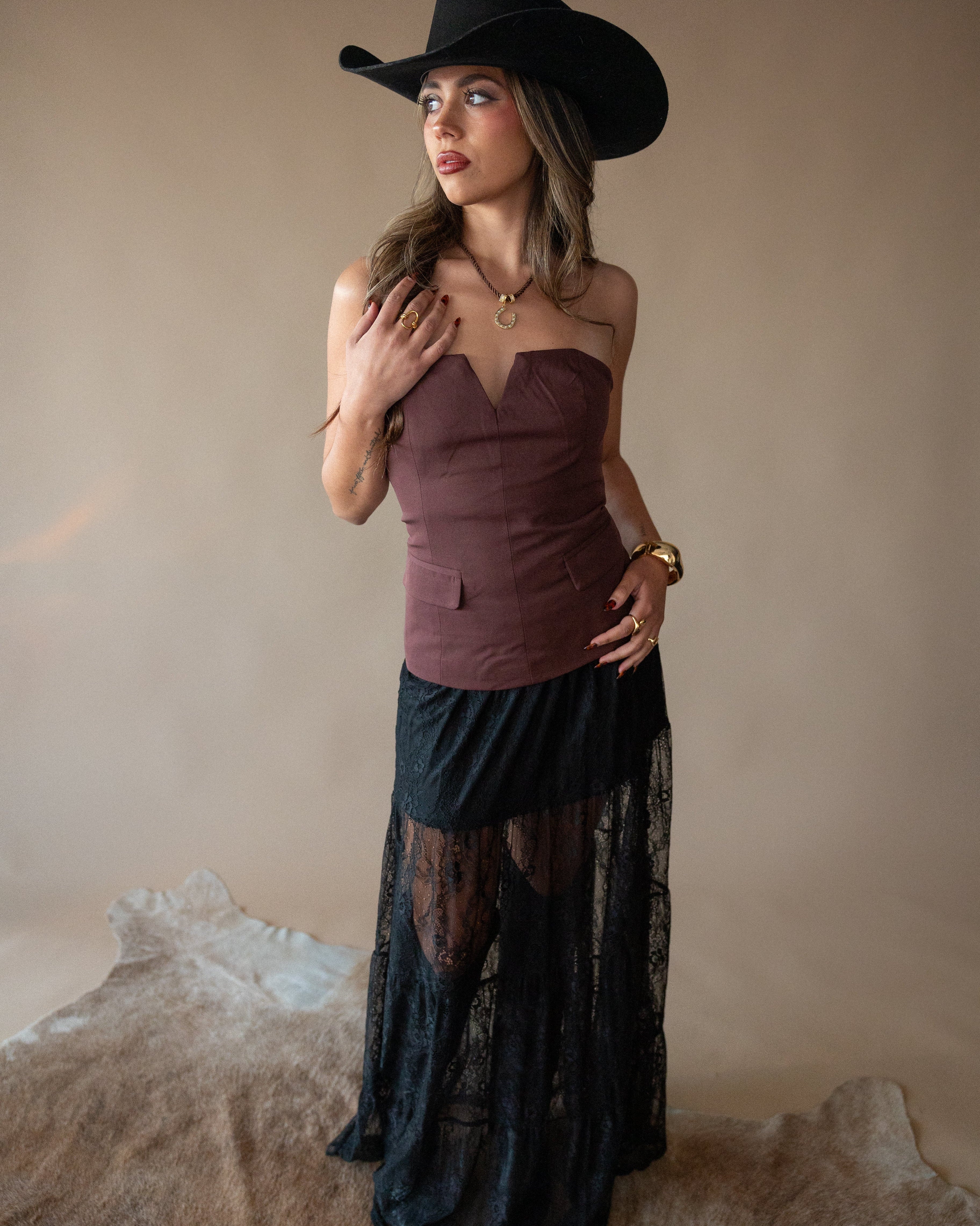 Woman wearing a purple top and black lace skirt with a black cowboy hat against a beige background