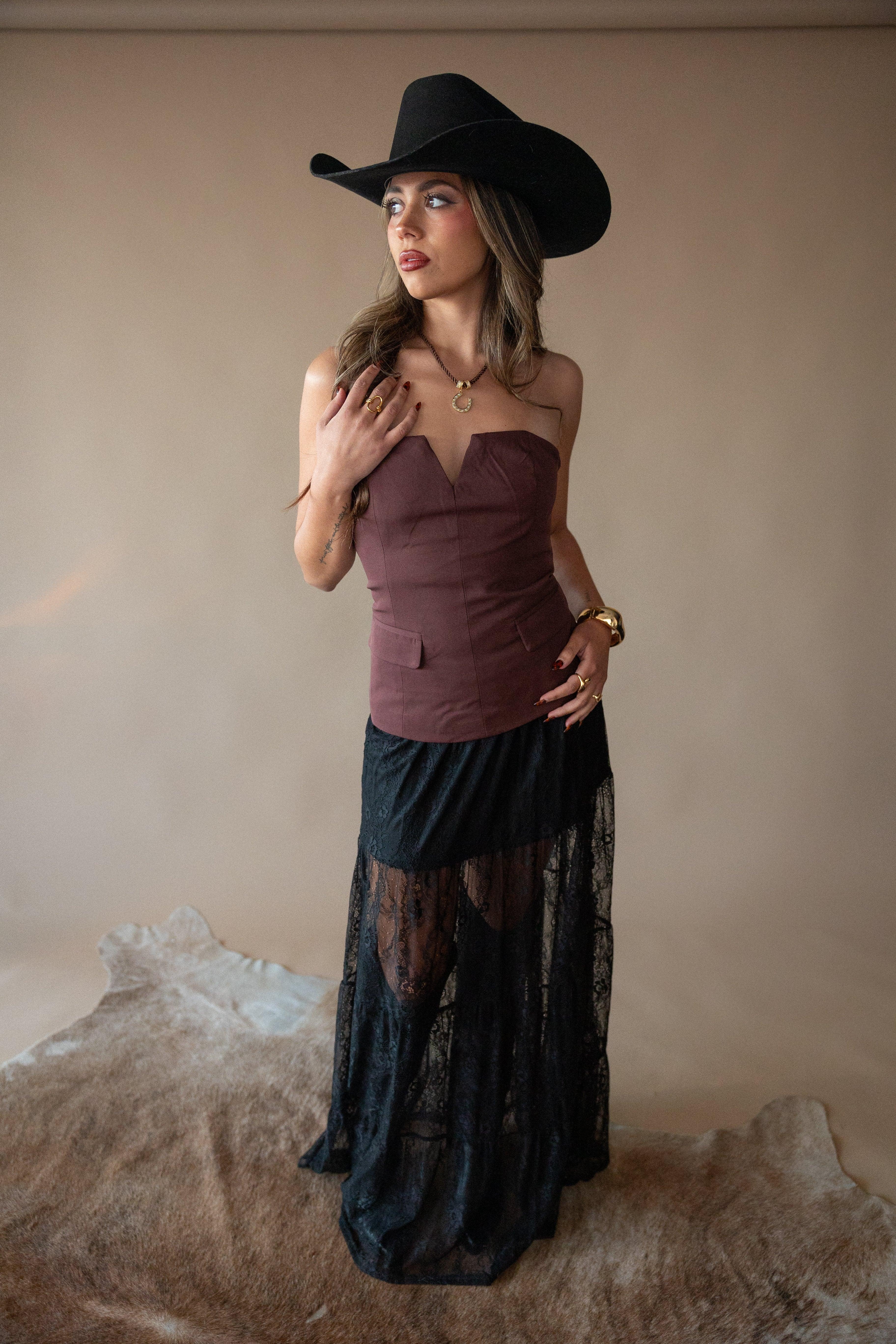 Woman wearing a purple top and black lace skirt with a black cowboy hat against a beige background