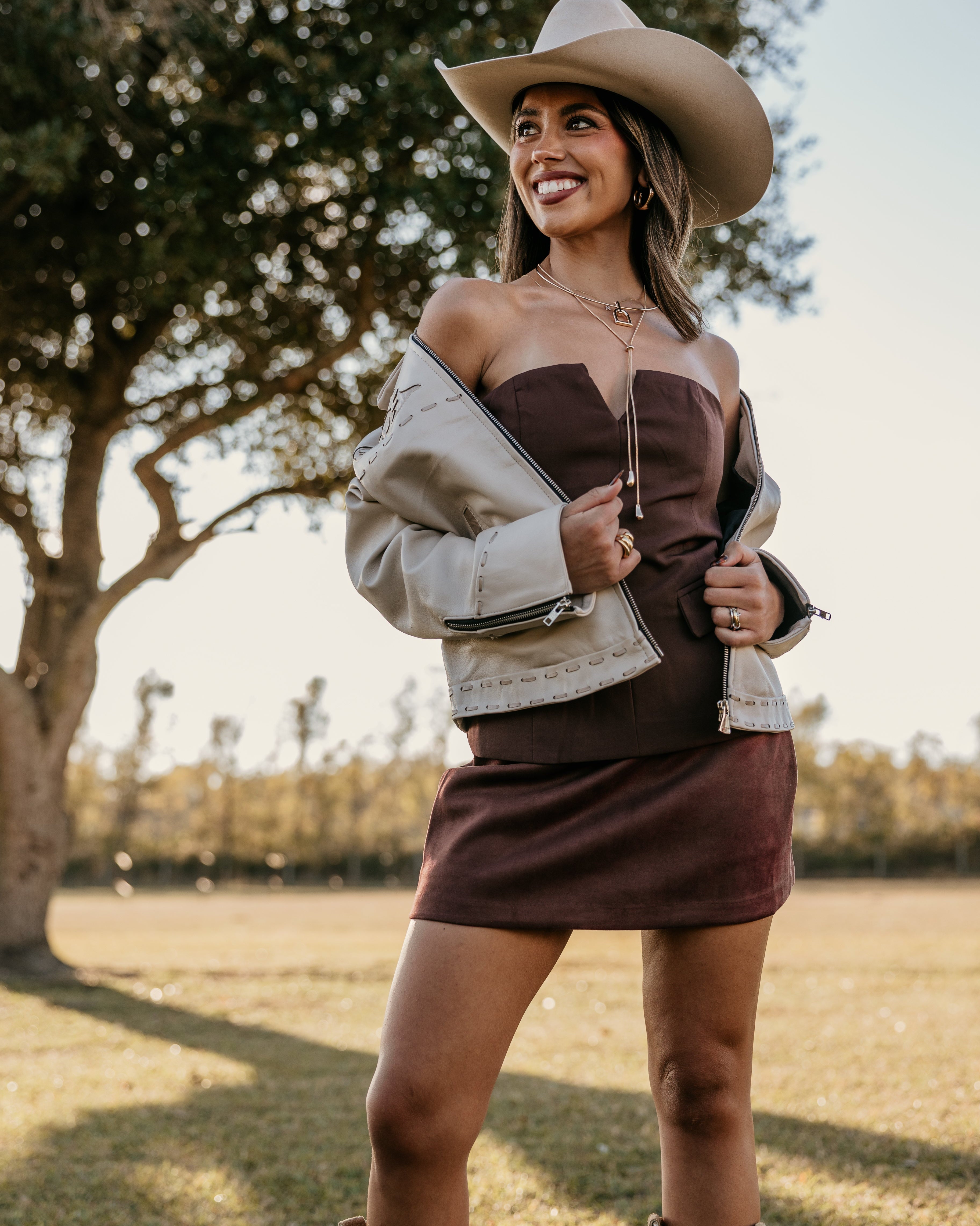 Woman wearing a cowboy hat, off-shoulder sweater, and skirt in an outdoor setting.