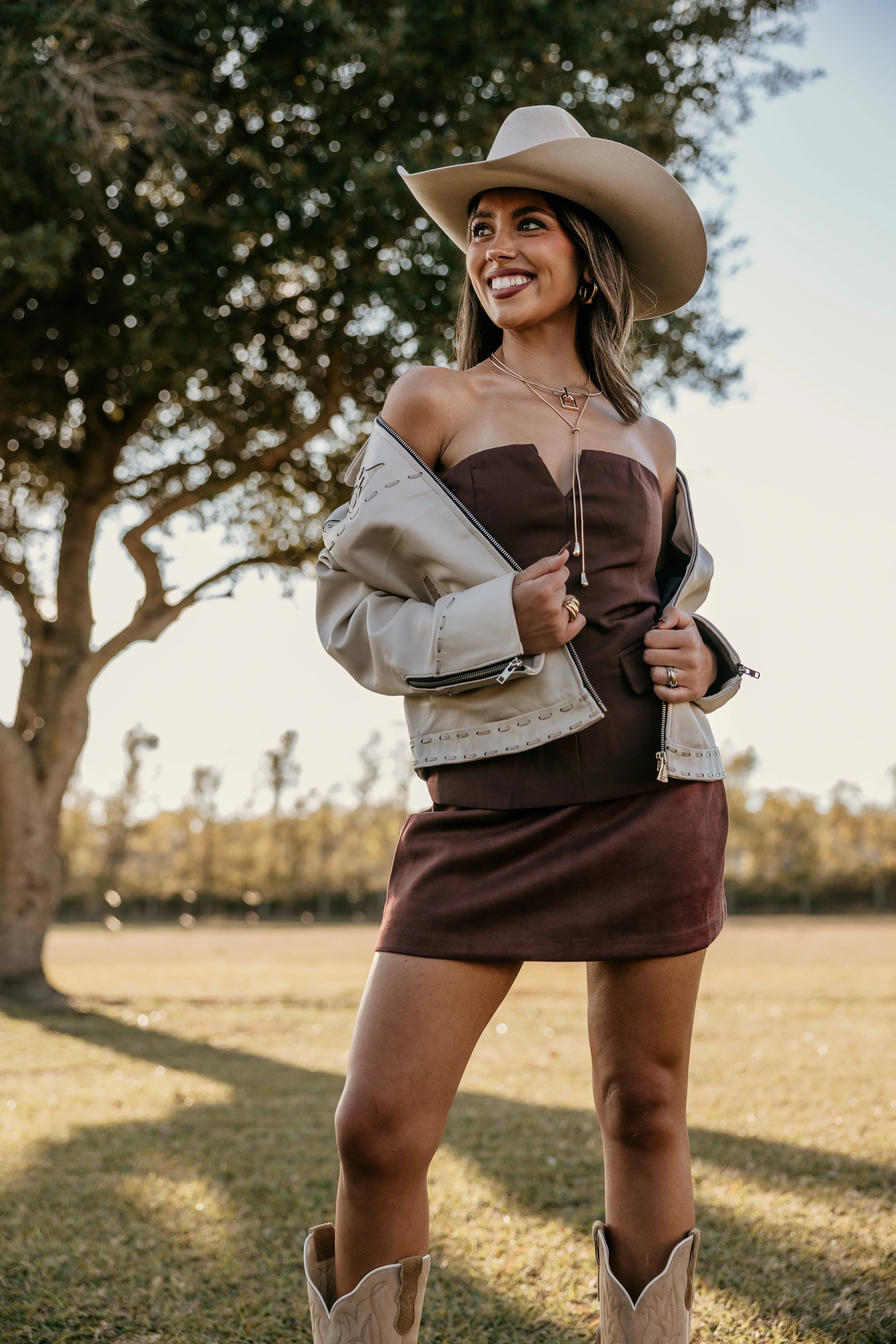 Woman wearing a cowboy hat, off-shoulder sweater, and skirt in an outdoor setting.