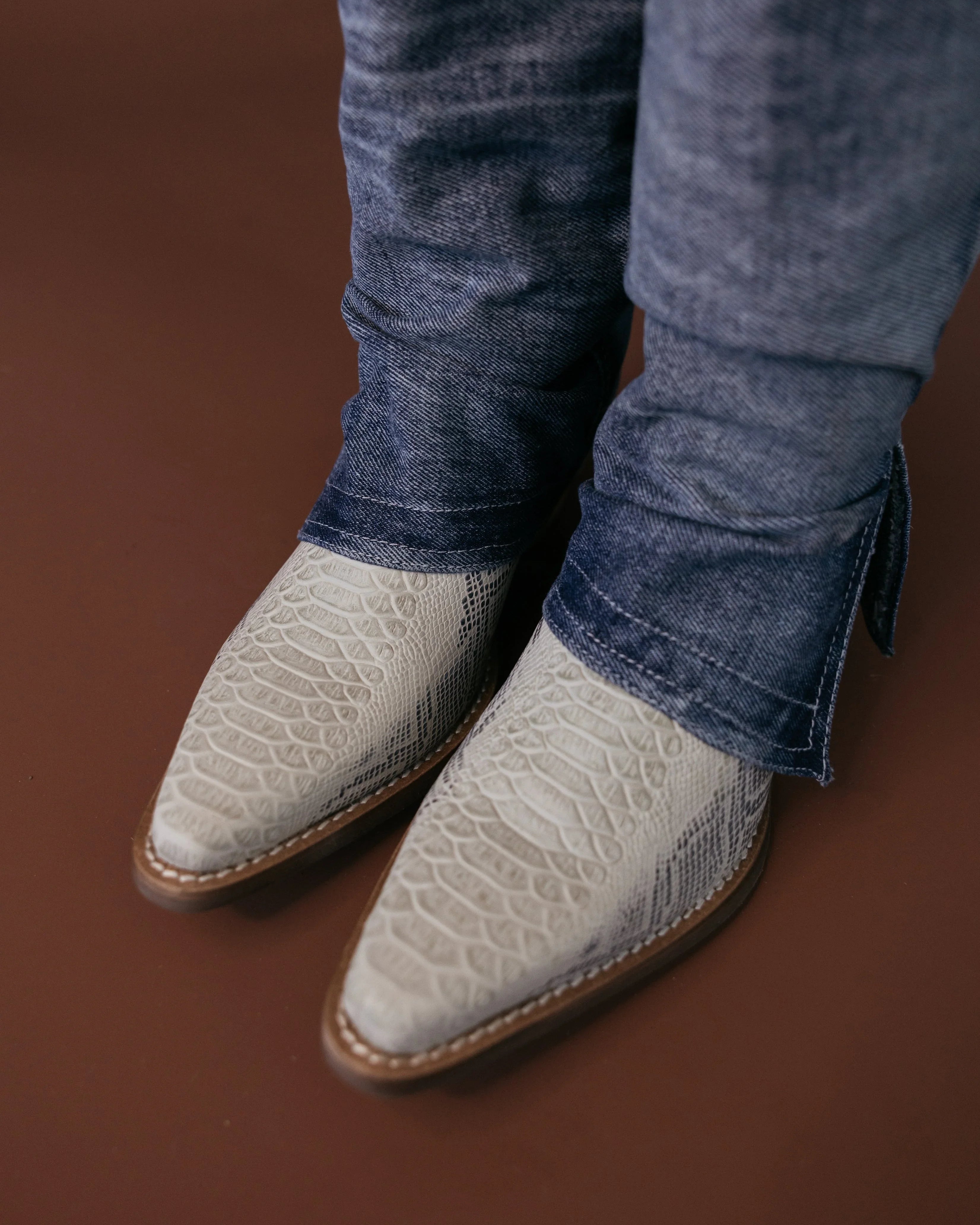 White textured western cowboy boots under blue jeans on a brown background