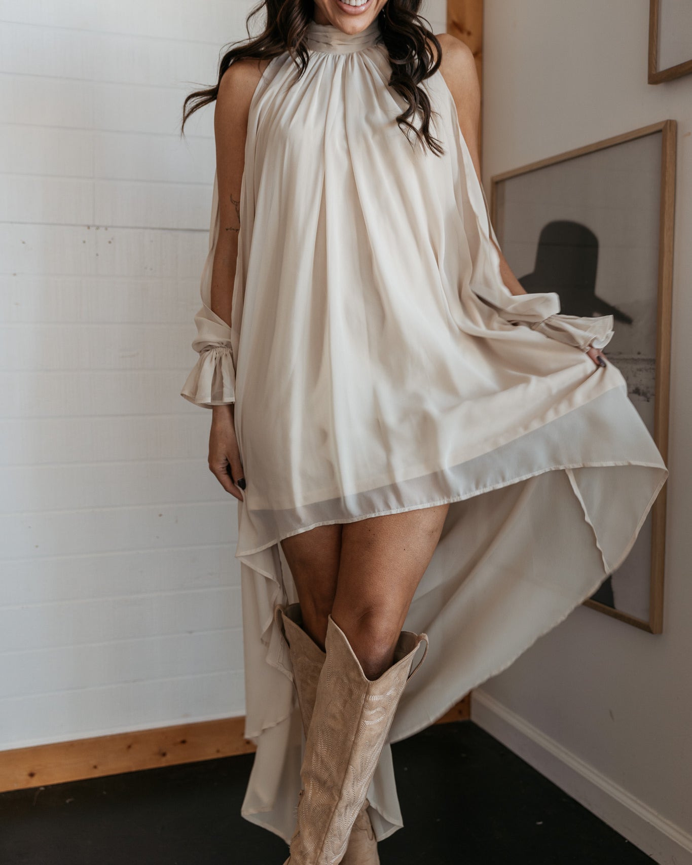 Woman in a white dress and beige boots standing indoors with a white wall and framed picture in the background.