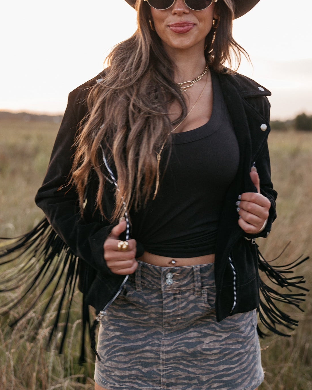 Woman wearing a black cowboy hat, fringe jacket, and sunglasses in a field.