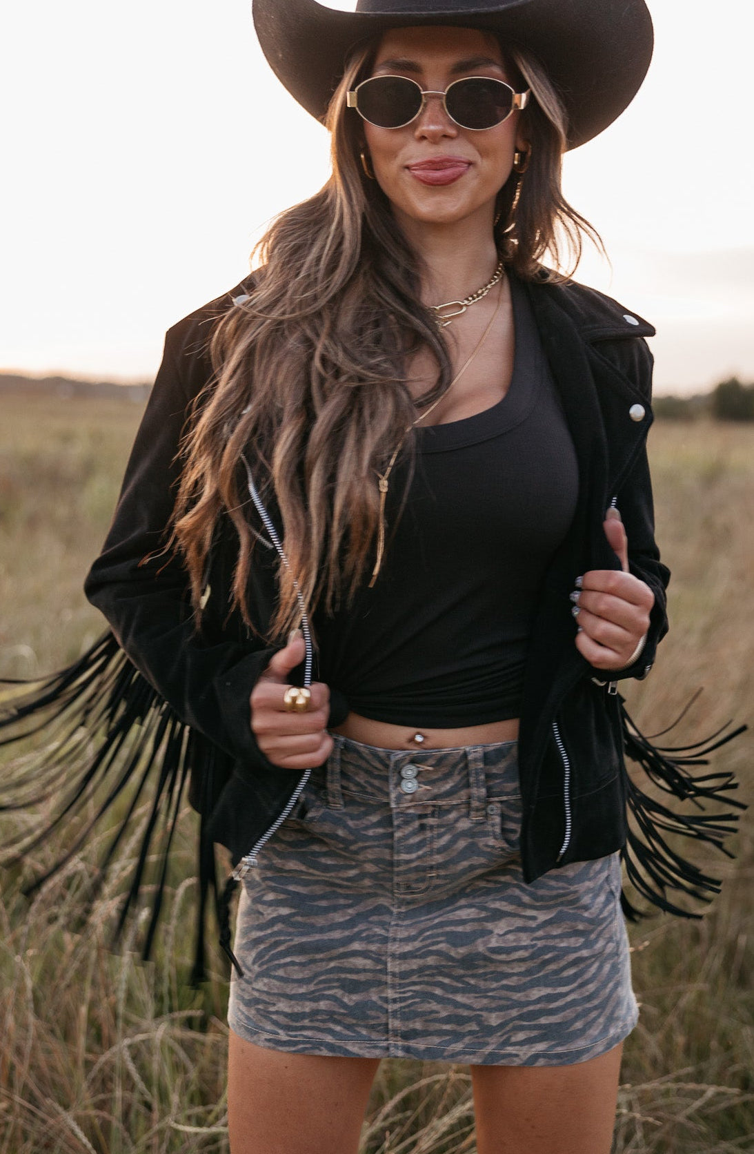 Woman wearing a black cowboy hat, fringe jacket, and sunglasses in a field.