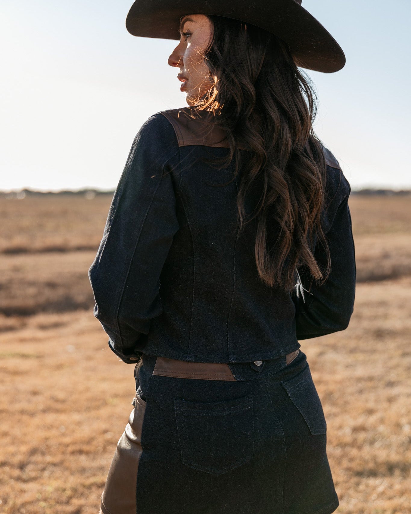 Woman in western denim jacket, mini skirt, and cowboy hat standing outdoors in a field