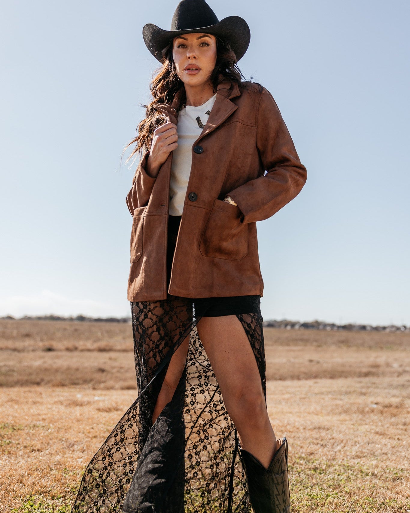 Woman in western outfit with brown suede jacket, black lace skirt, cowboy hat and boots outdoors