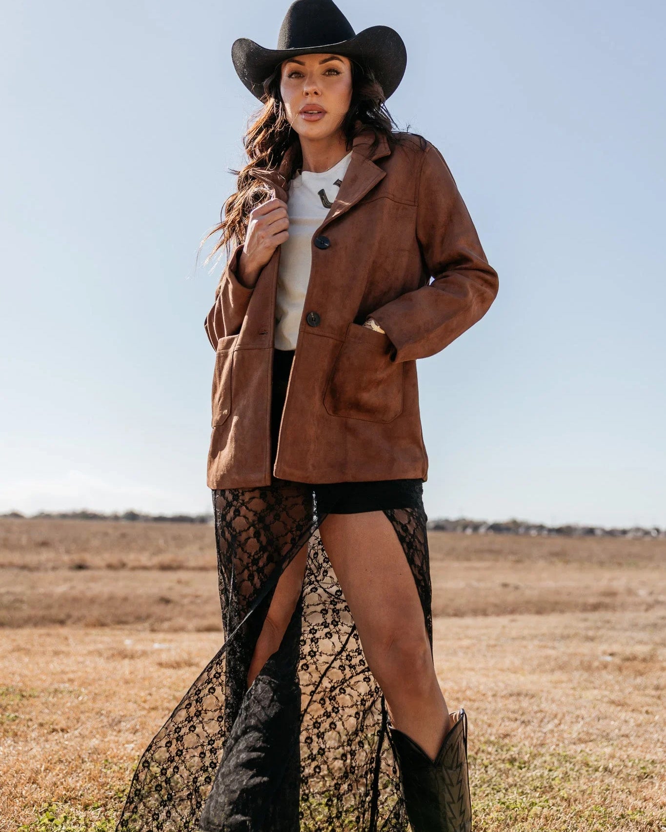 Woman in western outfit with brown suede jacket, black lace skirt, cowboy hat and boots outdoors