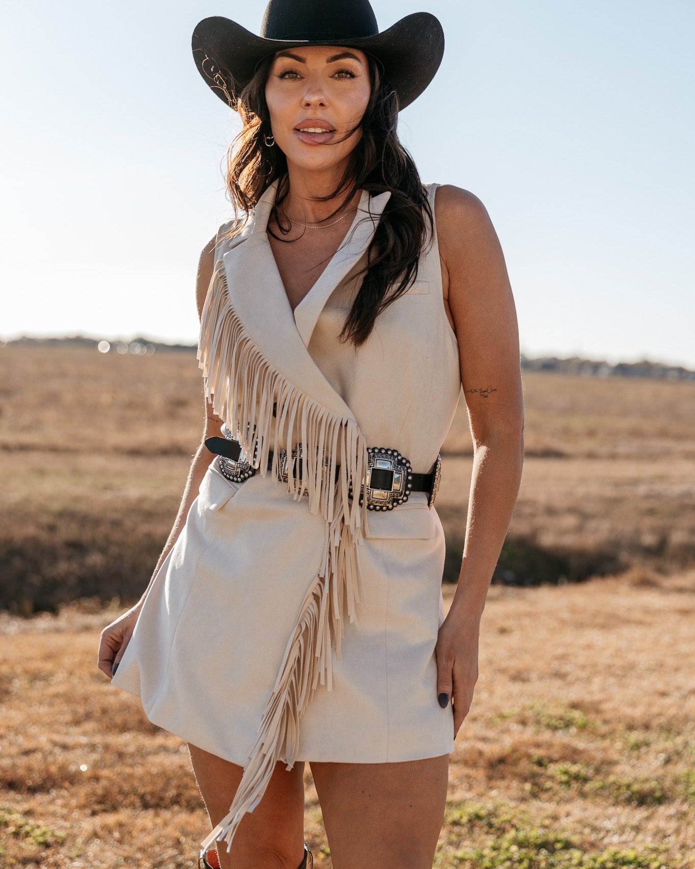 Woman in beige fringe western dress, black cowboy hat and boots outdoors, boho cowgirl style