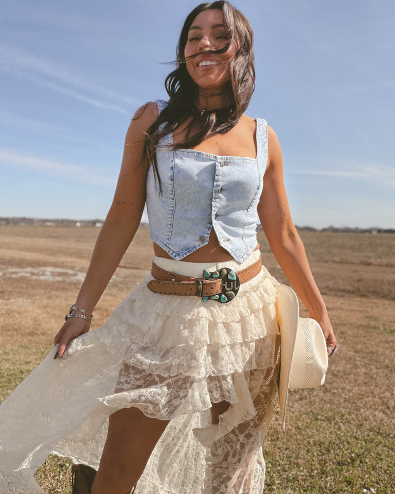 Woman in denim vest and lace skirt with western belt, holding cowboy hat outdoors