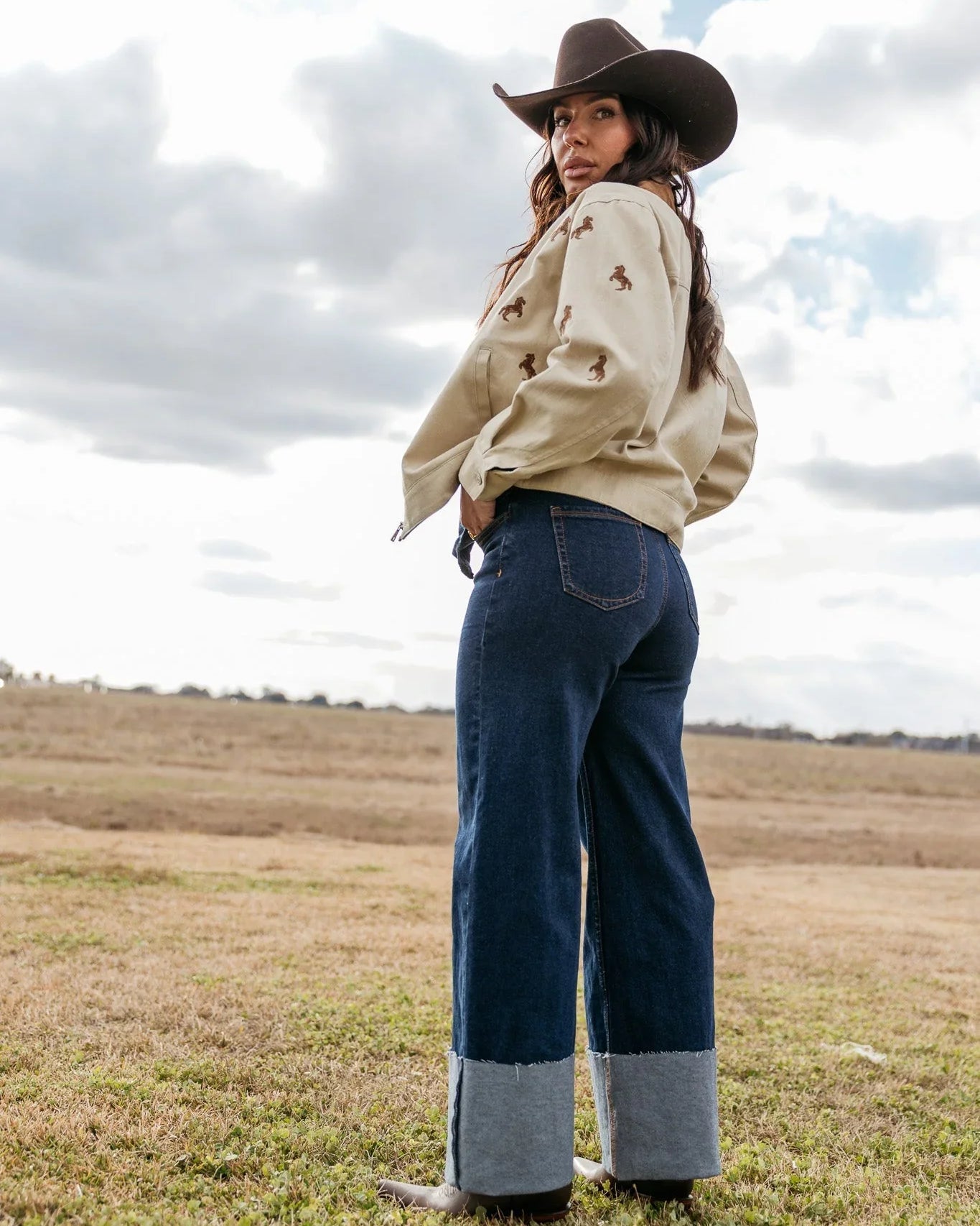 Woman in wide-leg denim jeans, beige horse print jacket, and cowboy hat in a western field