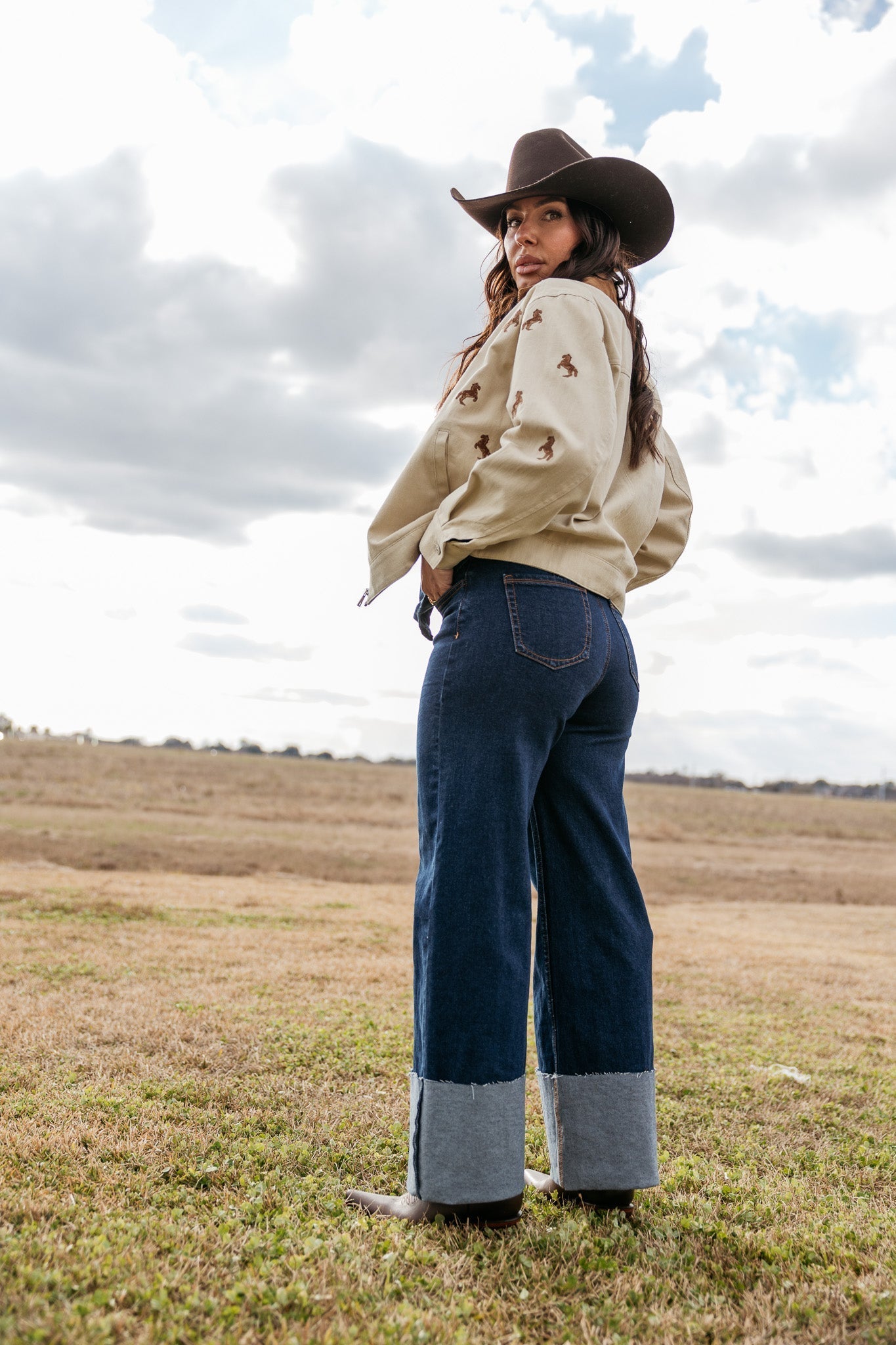 Woman in wide-leg denim jeans, beige horse print jacket, and cowboy hat in a western field