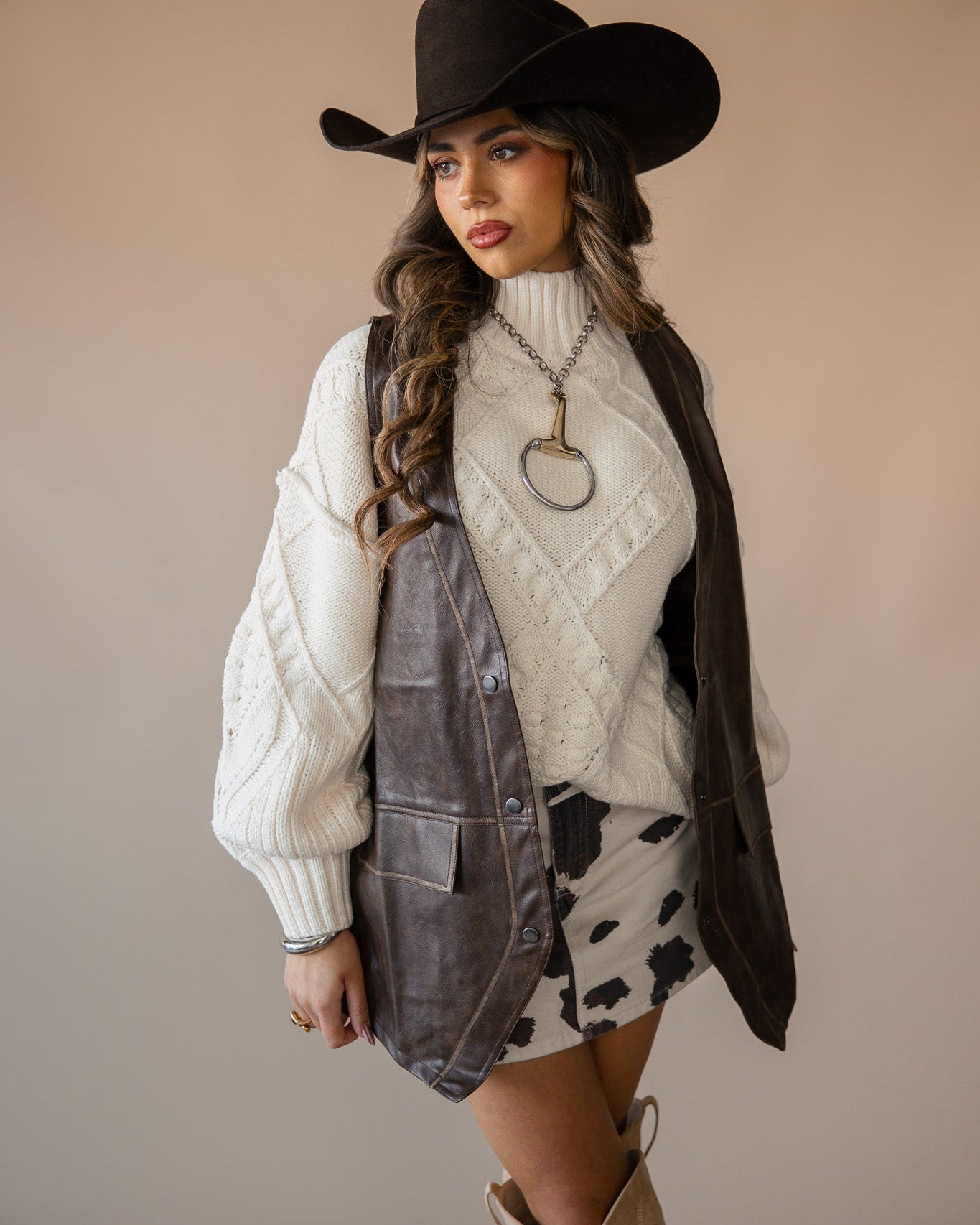 Woman in cowgirl hat, white sweater, brown vest, cow print skirt, and cowboy boots