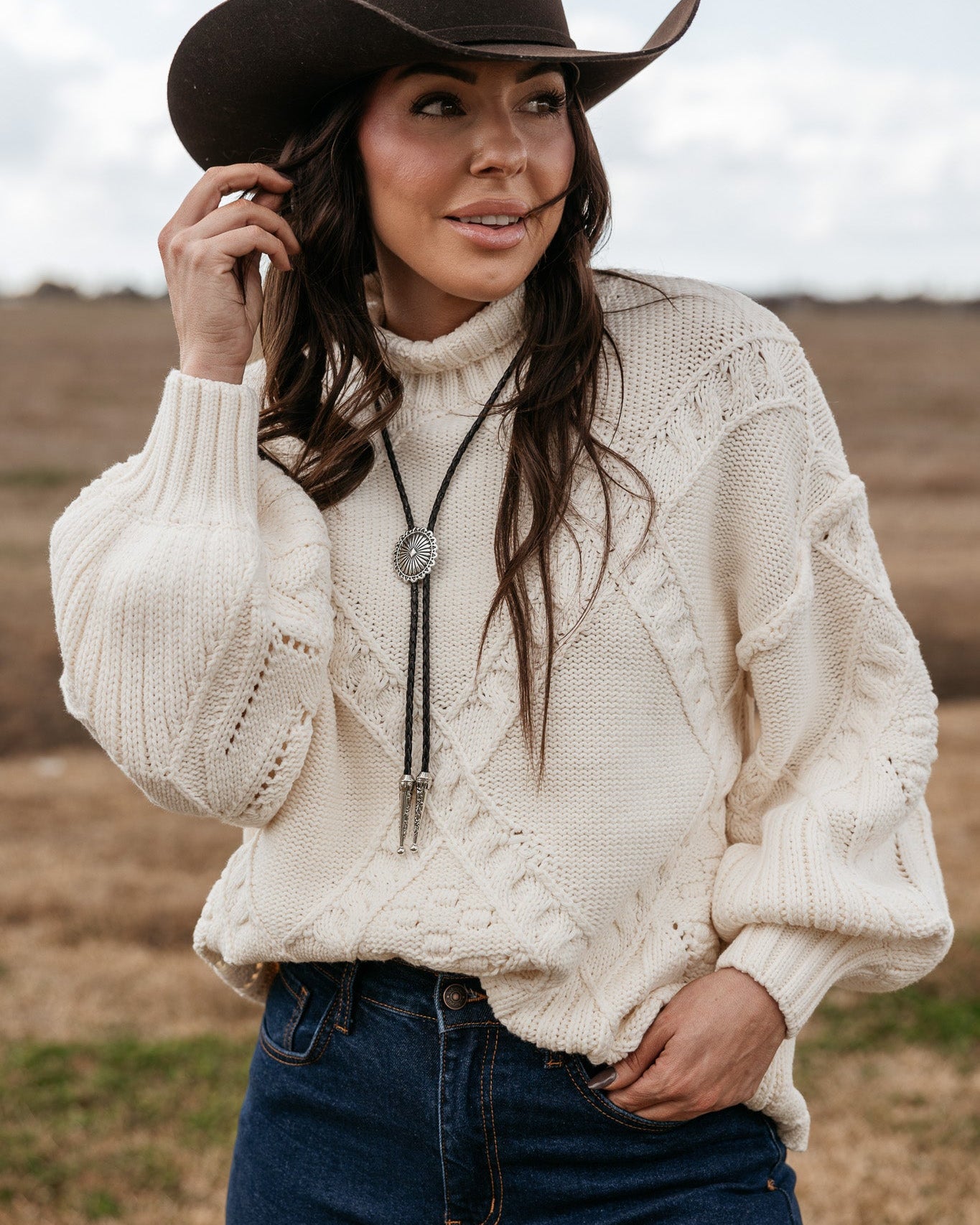 Woman in a brown cowboy hat, cream knit sweater, bolo tie, and jeans in a western field