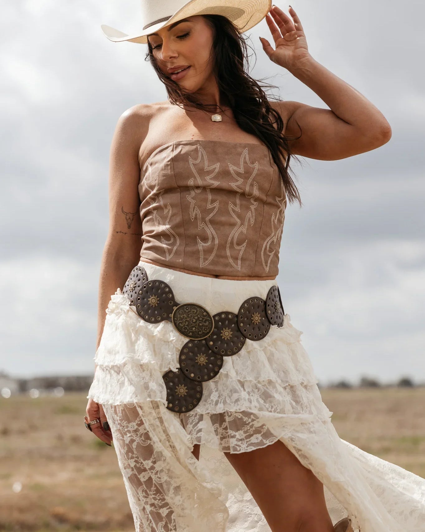 Woman in cowgirl hat, embroidered corset top, lace skirt, and statement western belt outdoors