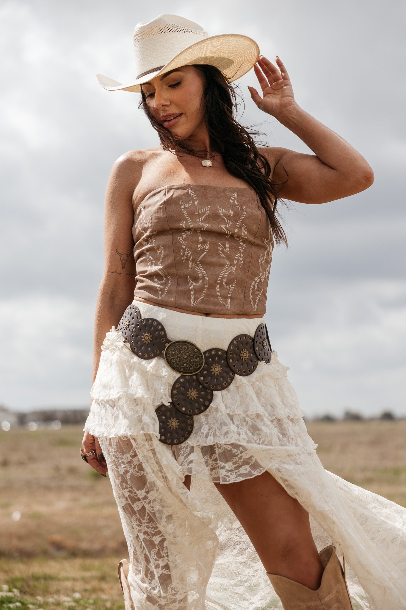 Woman in cowgirl hat, embroidered corset top, lace skirt, and statement western belt outdoors