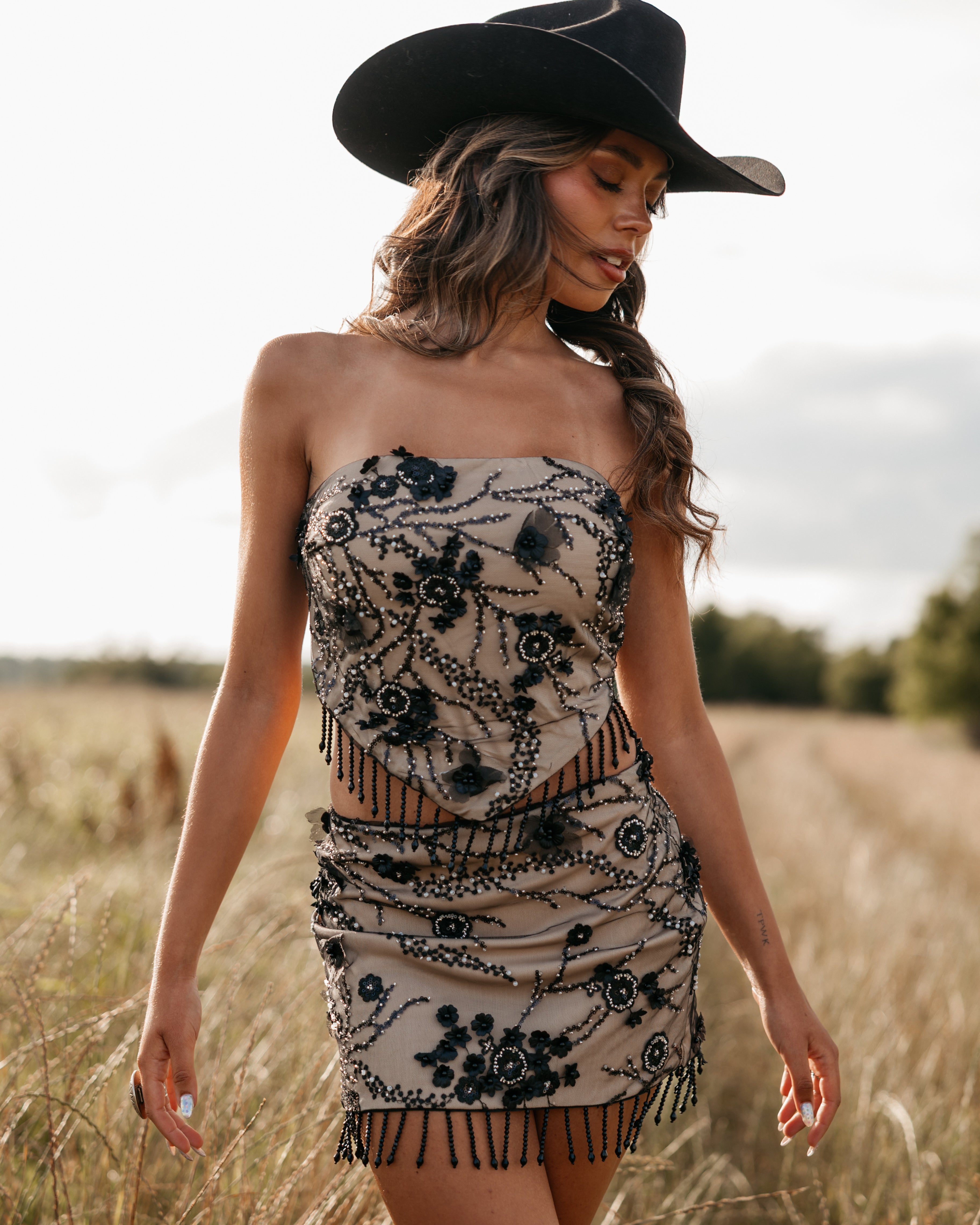 Woman in a strapless embroidered beaded set and black cowboy hat standing in a field.