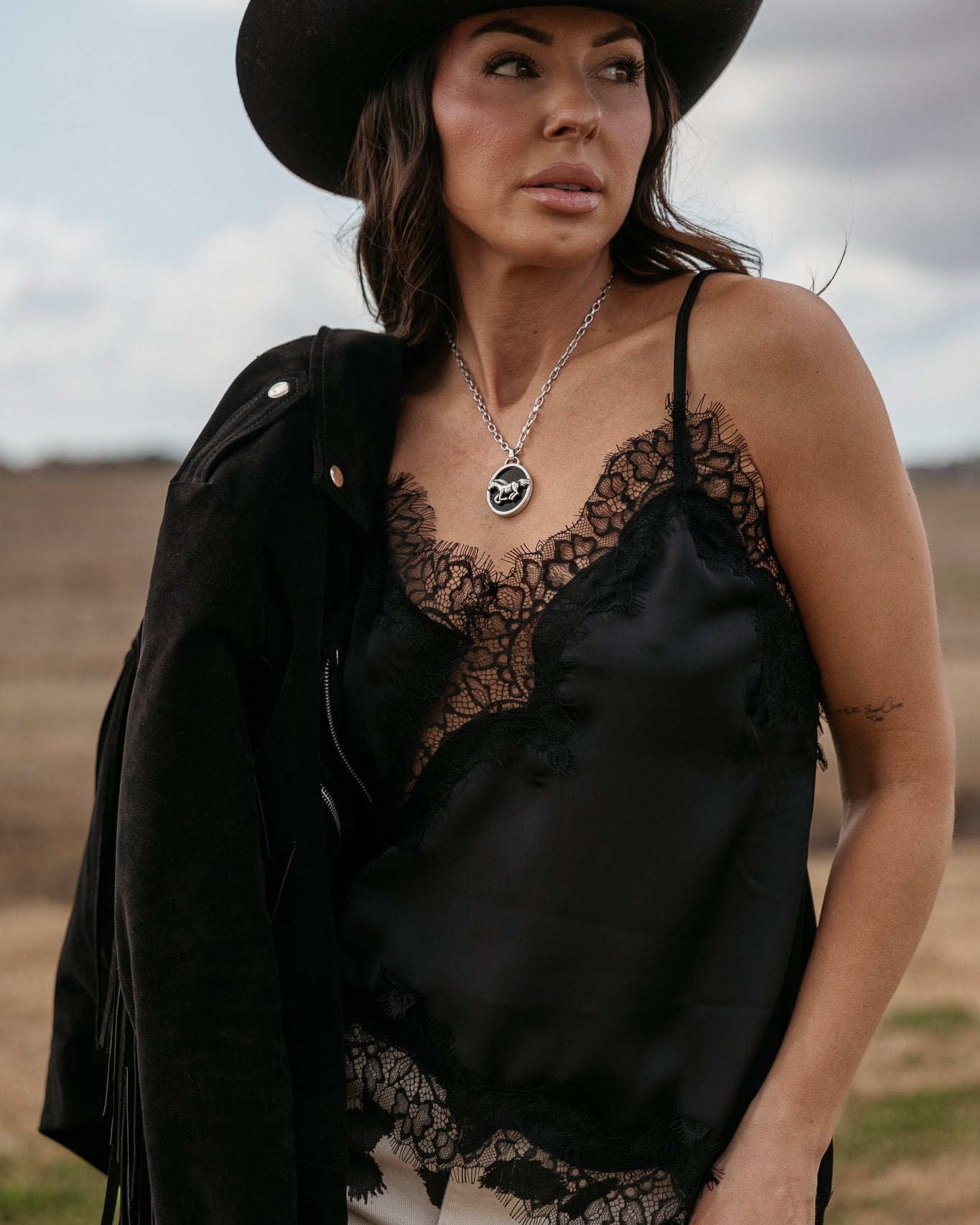 Woman in a black cowboy hat, black lace camisole, suede fringe jacket, and cow print pants outdoors