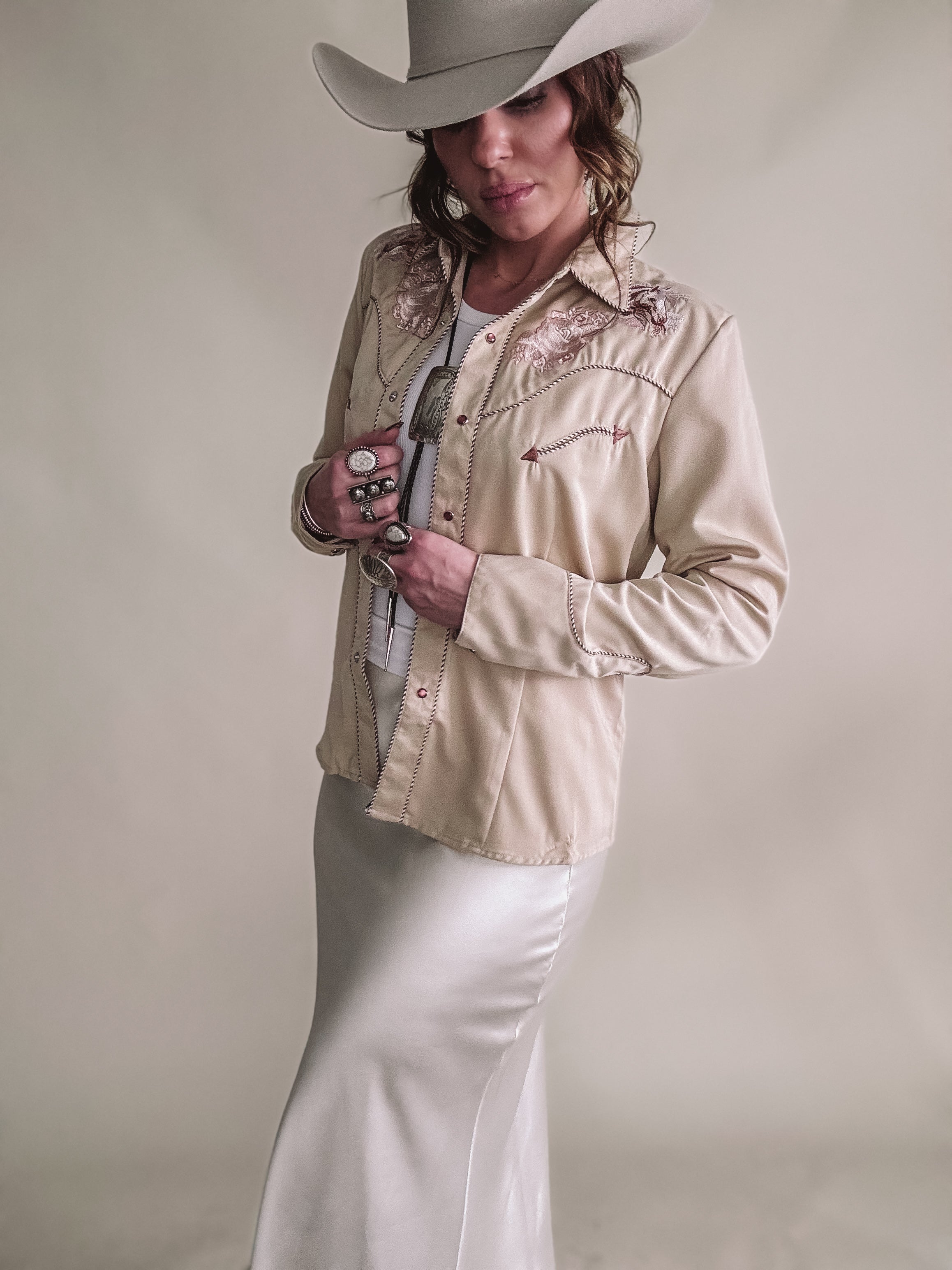 Woman in a cream western shirt, satin skirt, and cowboy hat, styled with boho rings.