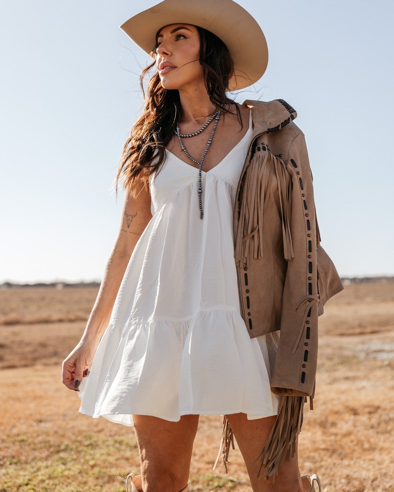 Woman in white western dress, cowboy hat, fringe suede jacket, and boots in outdoor setting