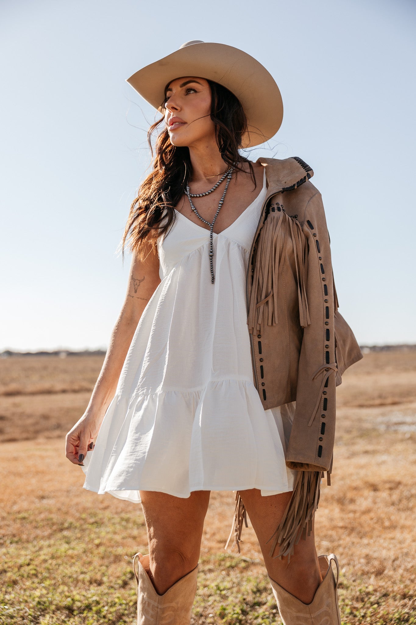 Woman in white western dress, cowboy hat, fringe suede jacket, and boots in outdoor setting