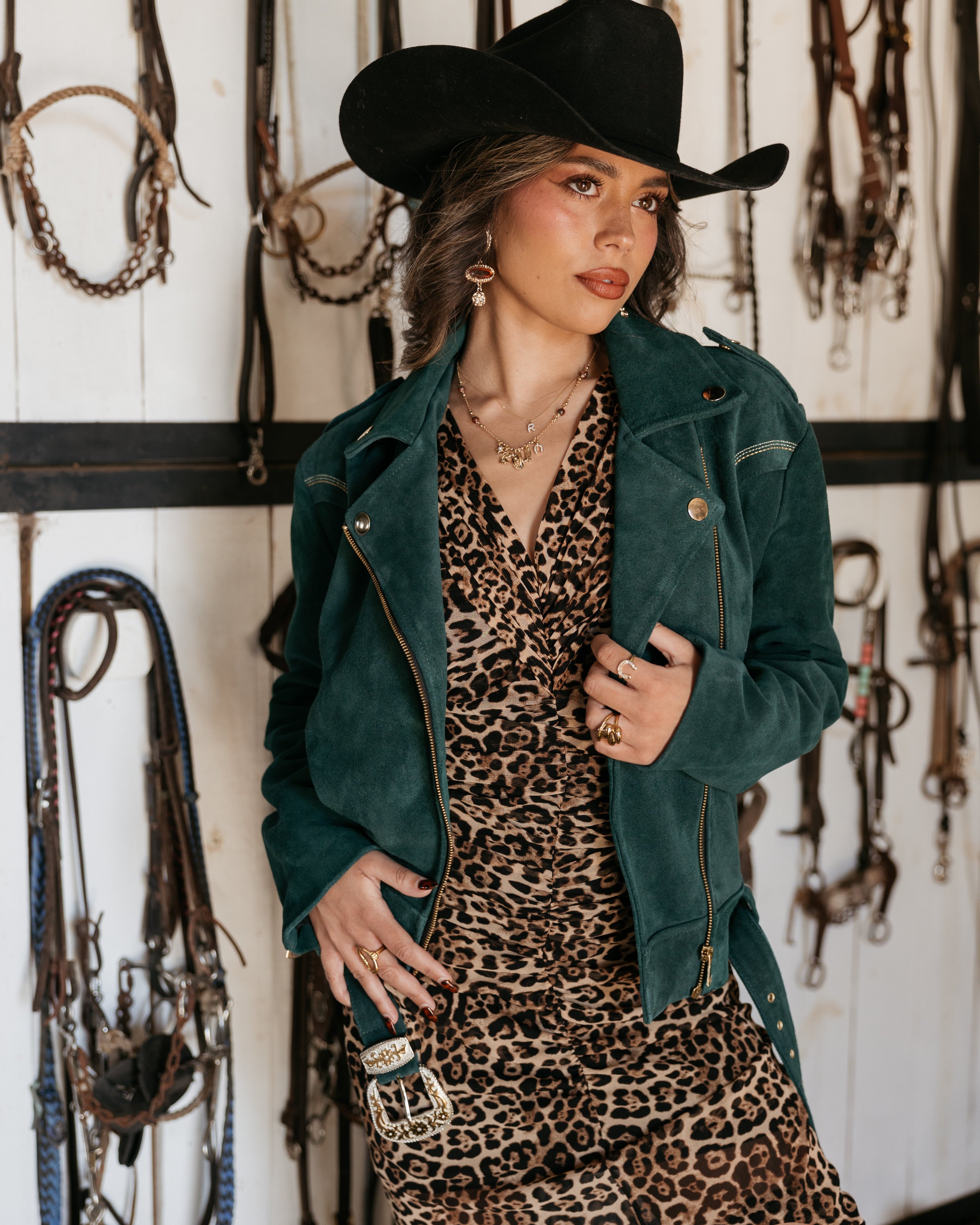 Woman in cowgirl hat, leopard print dress, and green suede jacket in western tack room