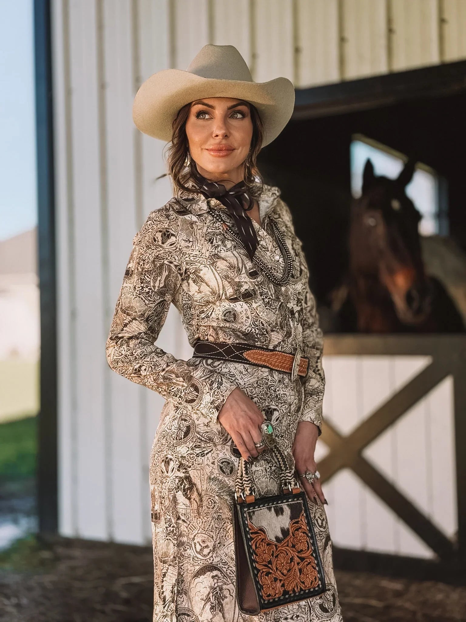 Woman in western print dress and cowboy hat with tooled leather purse, standing by horse stable