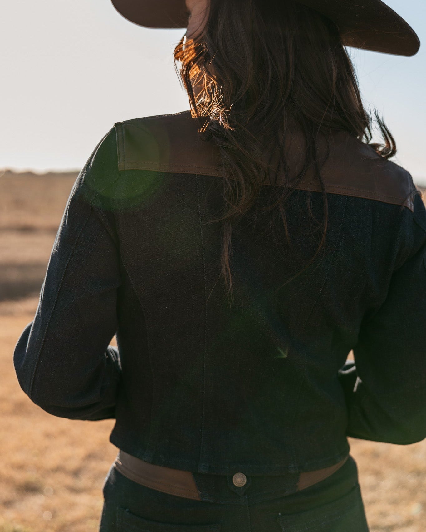 Woman in dark denim western jacket and cowboy hat, outdoors in sunlight, boho cowgirl style