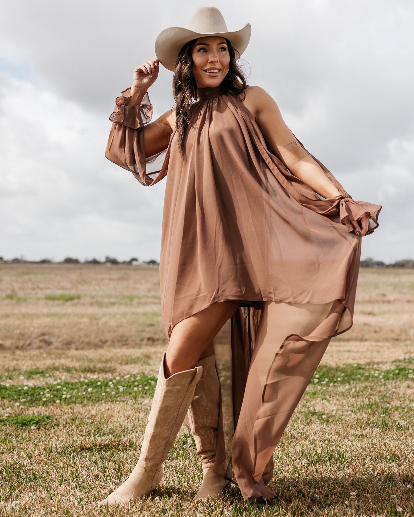 Woman in brown flowy western dress, ivory cowboy hat, and suede boots standing in a field