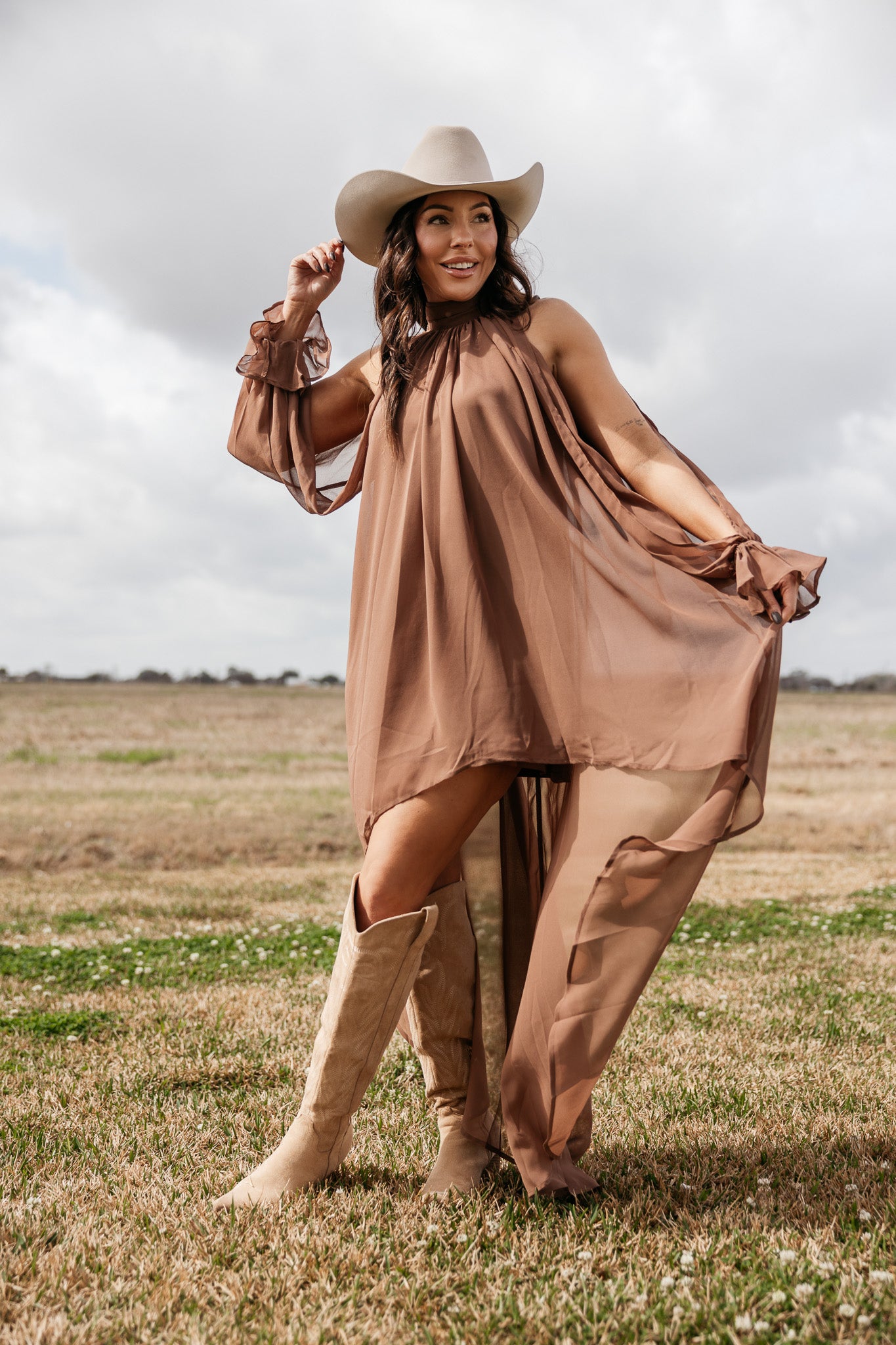 Woman in brown flowy western dress, ivory cowboy hat, and suede boots standing in a field