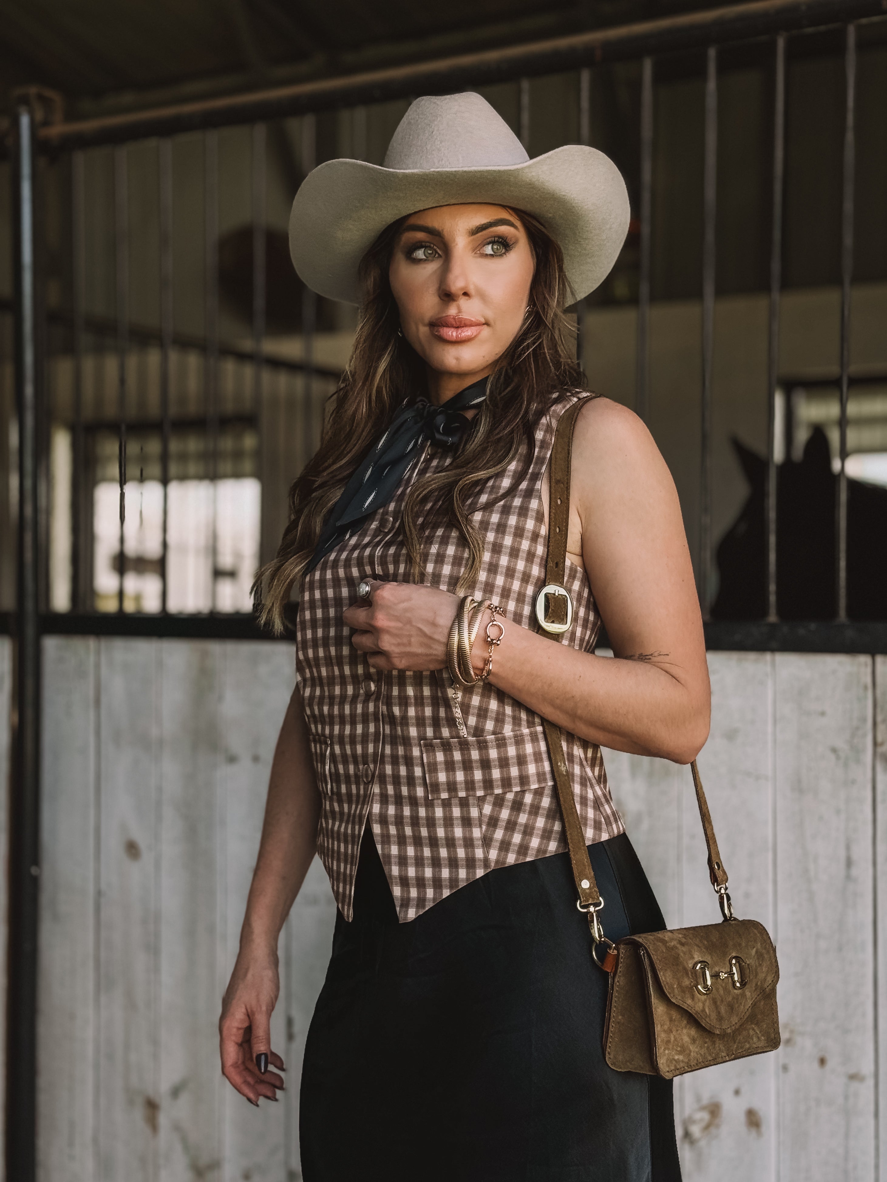 Woman in western cowgirl outfit with white hat, gingham vest, and suede purse in barn setting