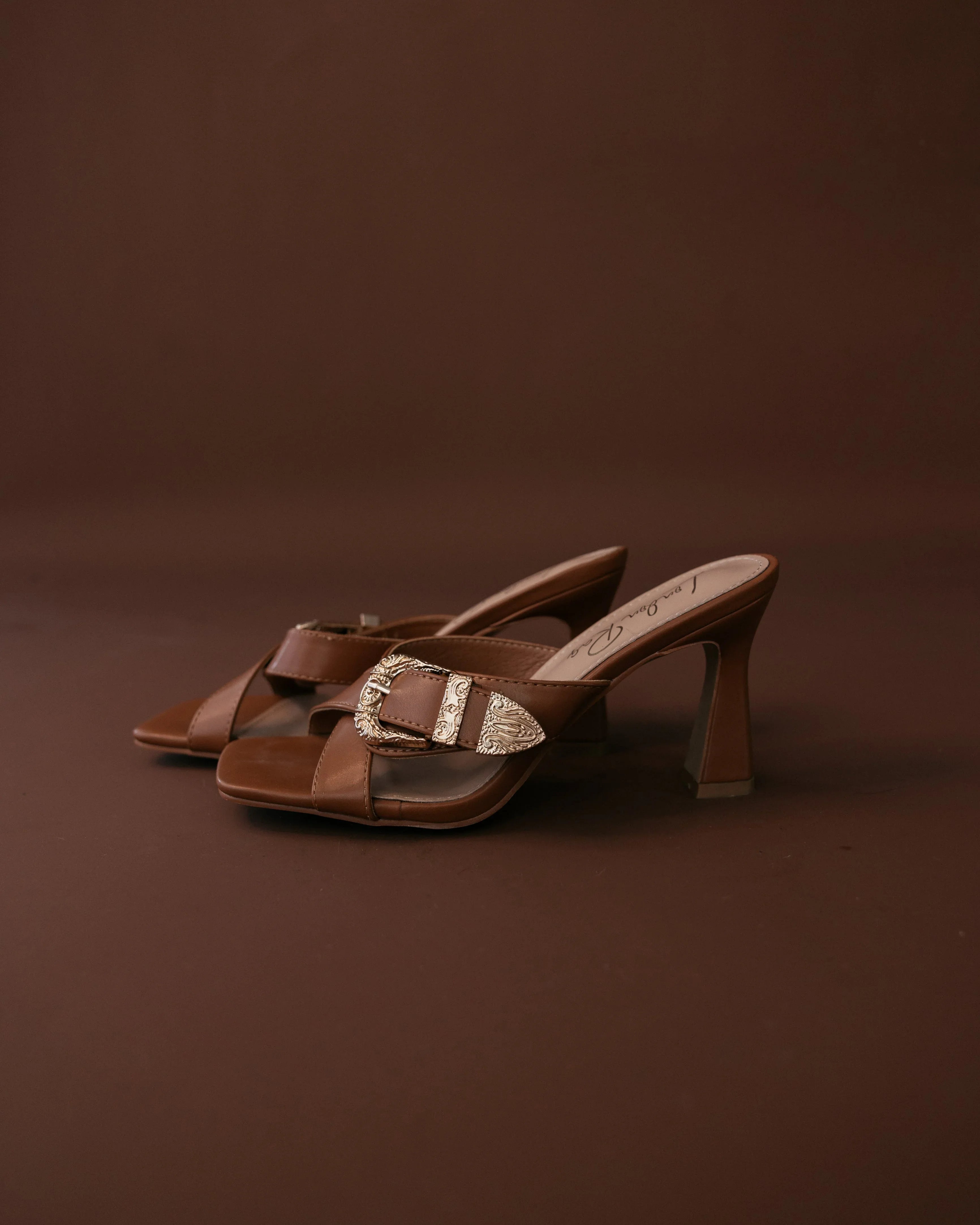 Brown western heeled sandals with gold buckle detail on brown background