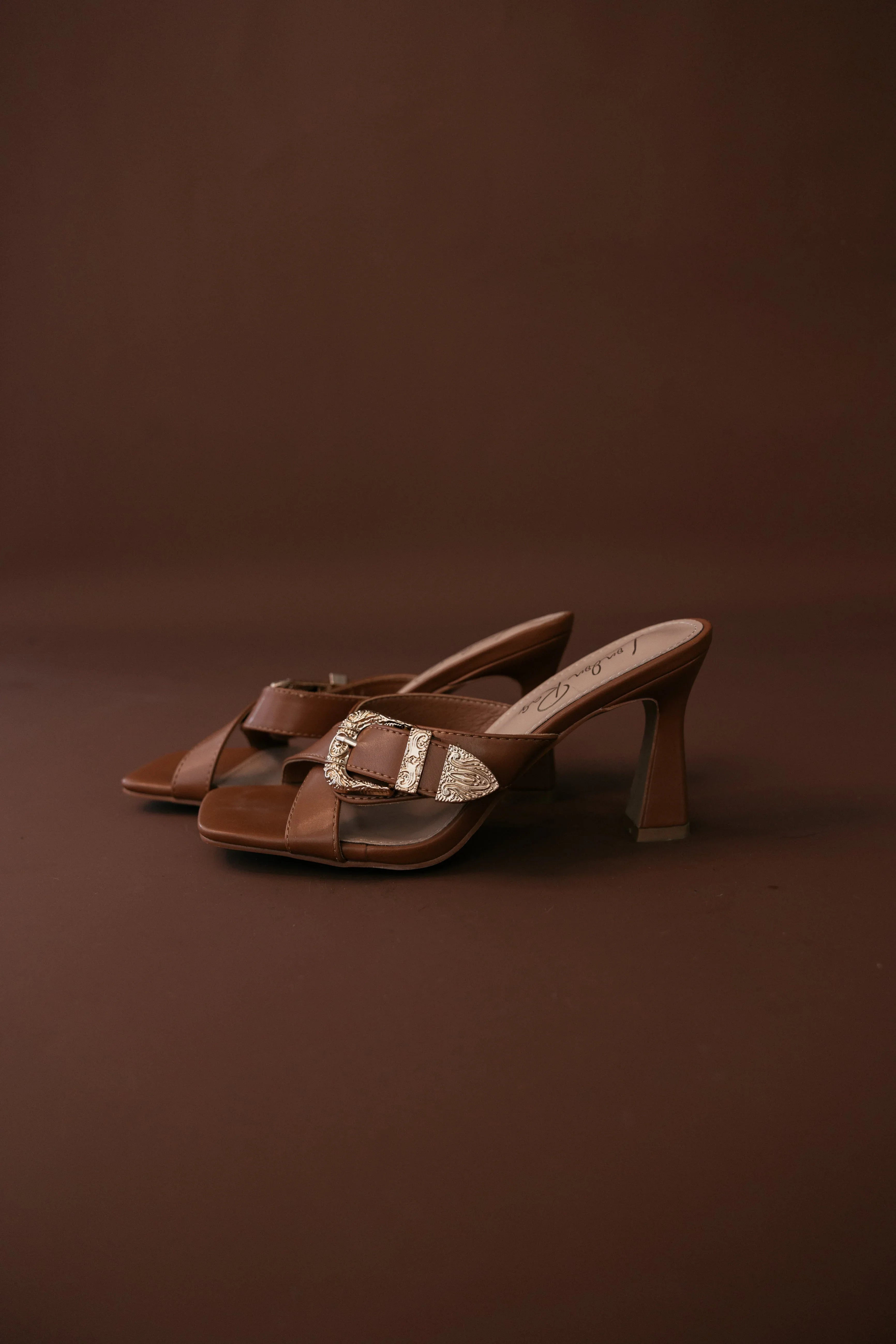 Brown western heeled sandals with gold buckle detail on brown background