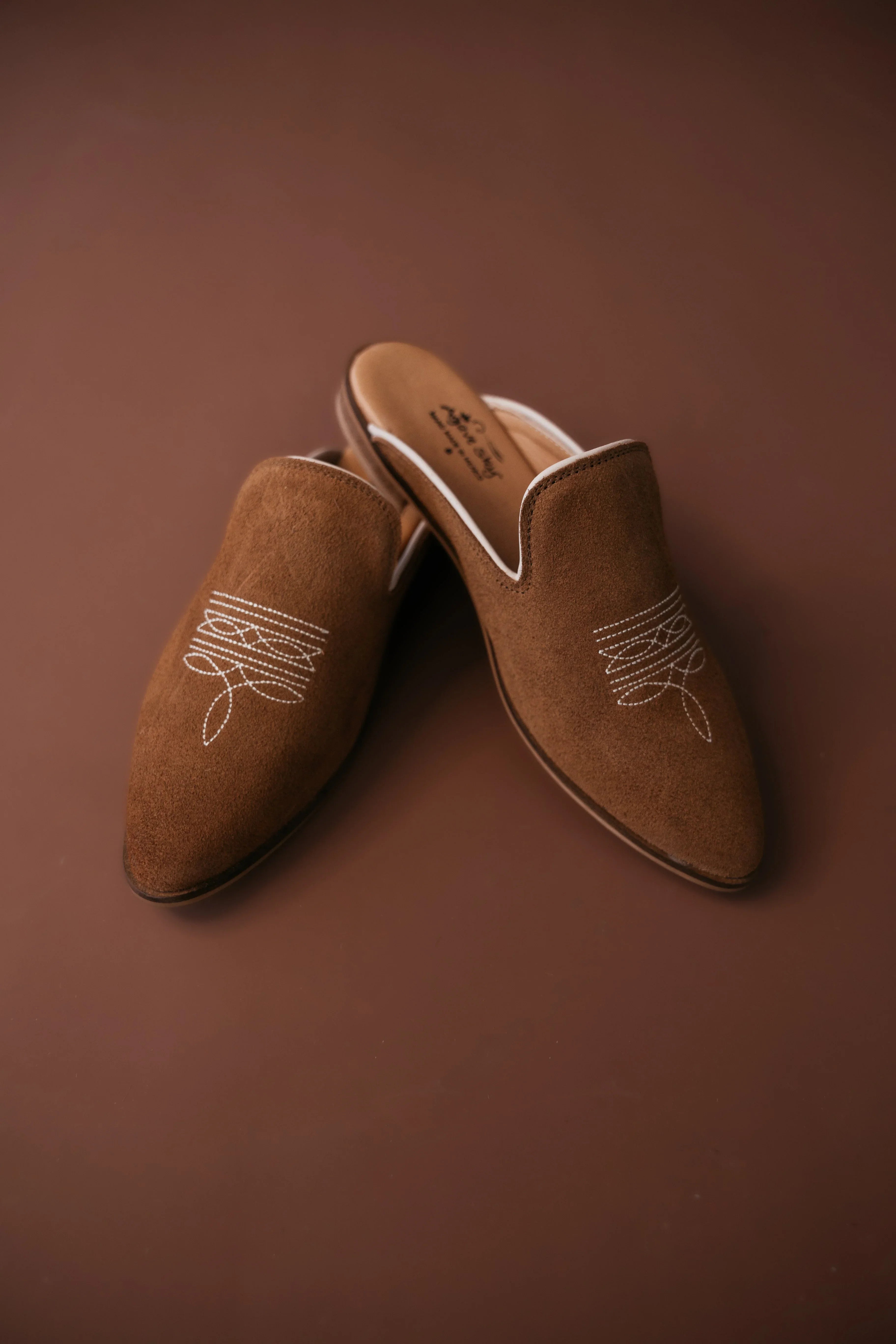 Pair of brown suede slippers with white embroidery on a brown background