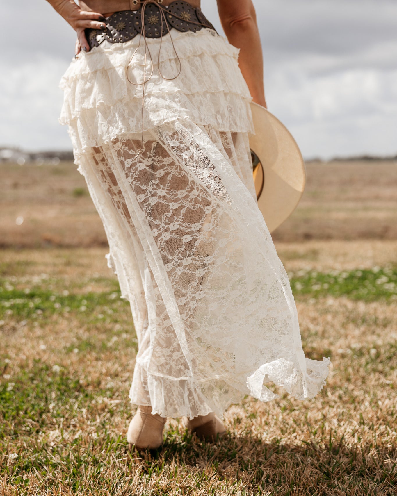 Woman in western lace maxi skirt, suede corset belt, and boots holding a cowboy hat outdoors