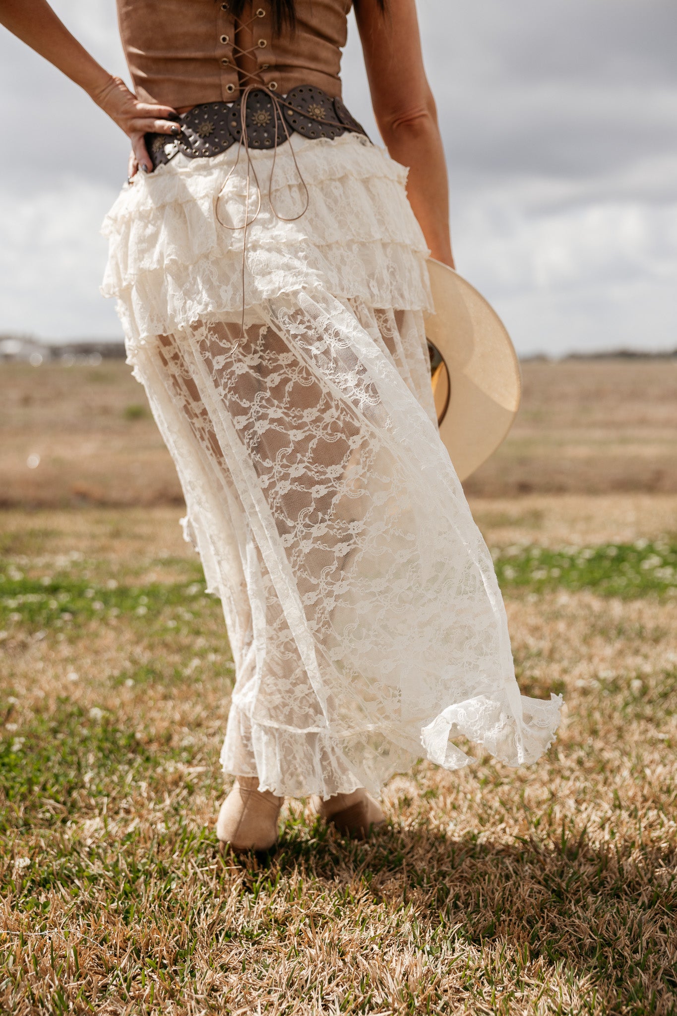 Woman in western lace maxi skirt, suede corset belt, and boots holding a cowboy hat outdoors