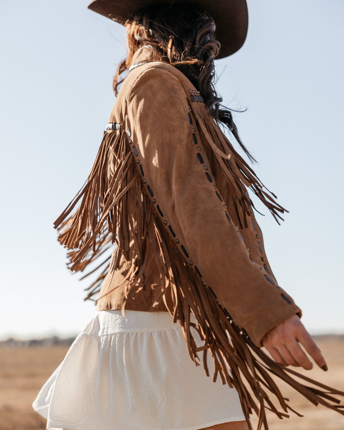 Woman in brown suede fringe jacket, white skirt, and western hat in a sunny outdoor field