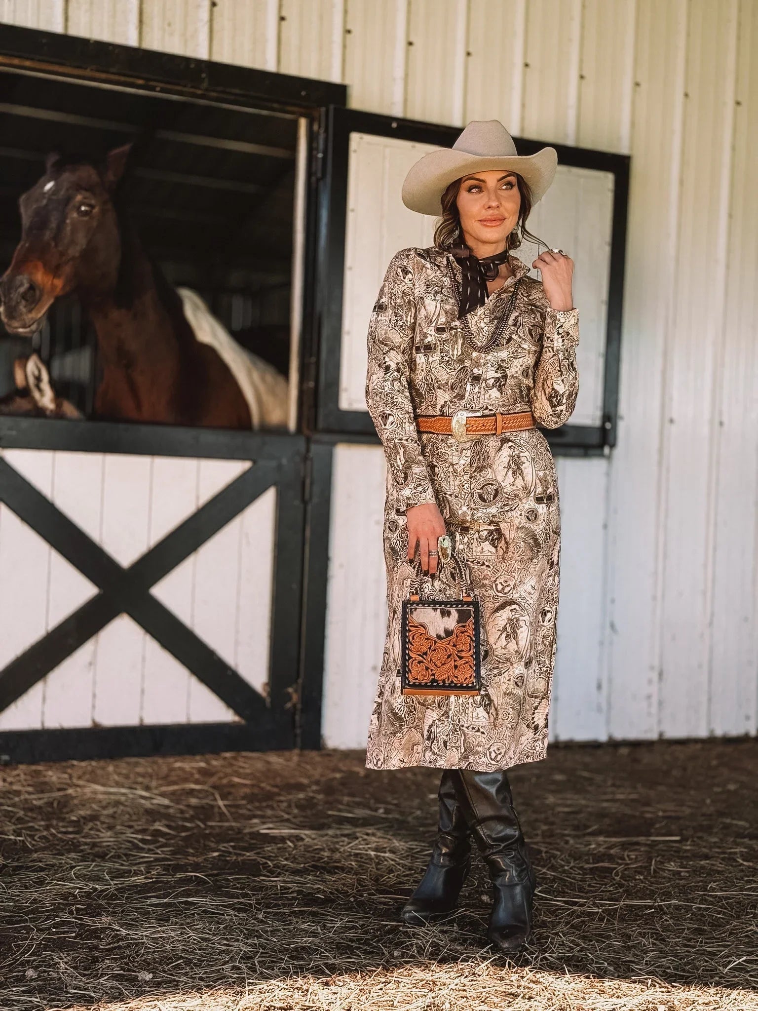 Woman in western boho dress, cowboy hat, and boots, standing by horse stable with horse.