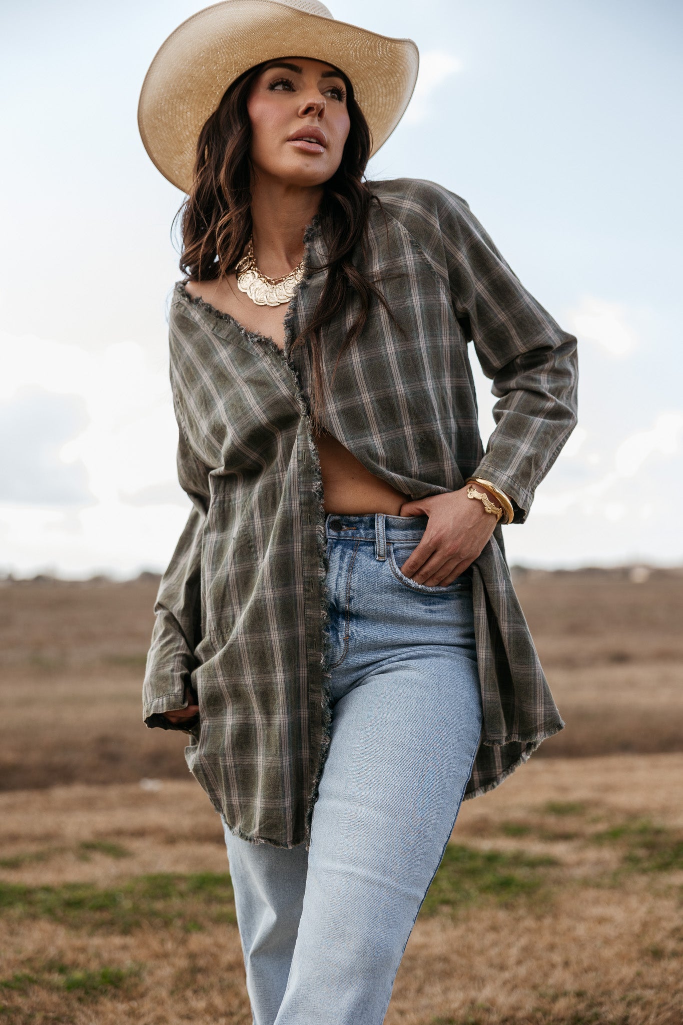 Woman in cowgirl hat, plaid western shirt, and light jeans in a rustic outdoor setting
