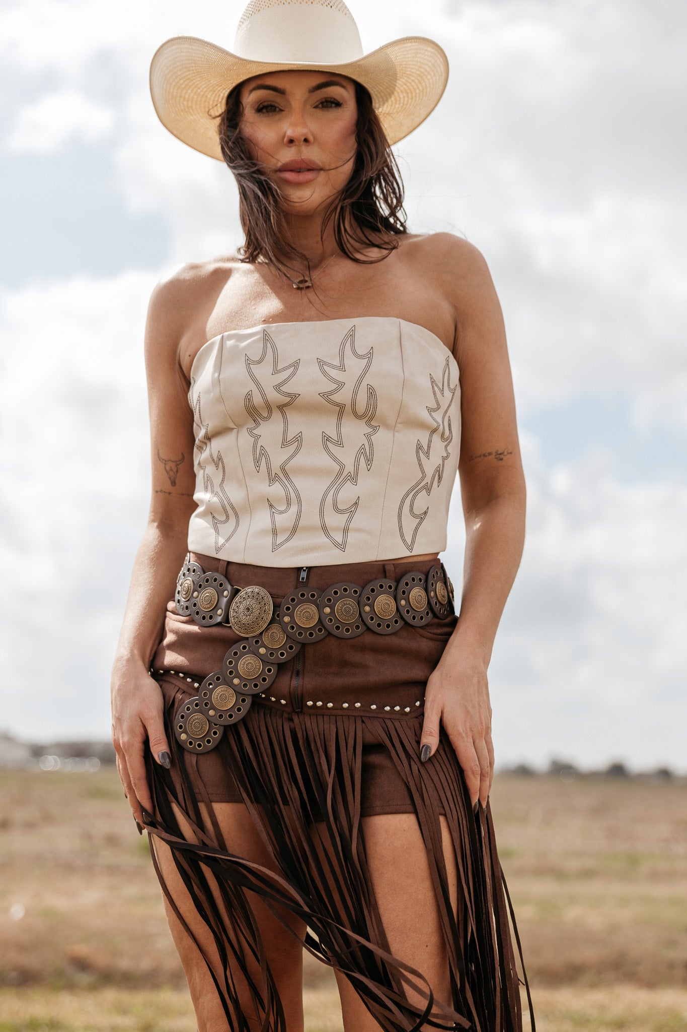 Woman in western boho outfit with white strapless top, fringed brown skirt, statement belt, and cowboy hat outdoors