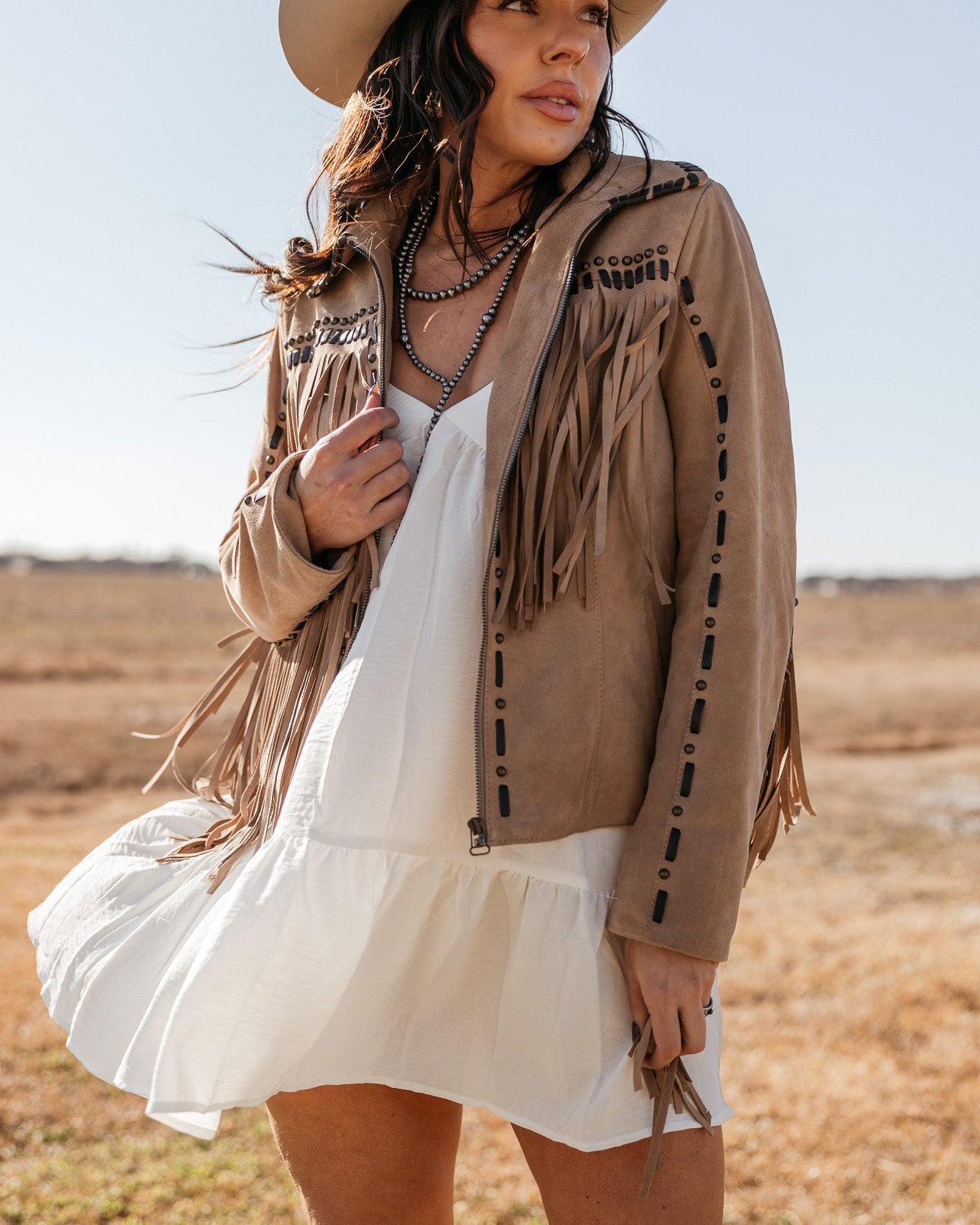 Woman in a tan fringe western jacket, white dress, and cowboy hat standing outdoors
