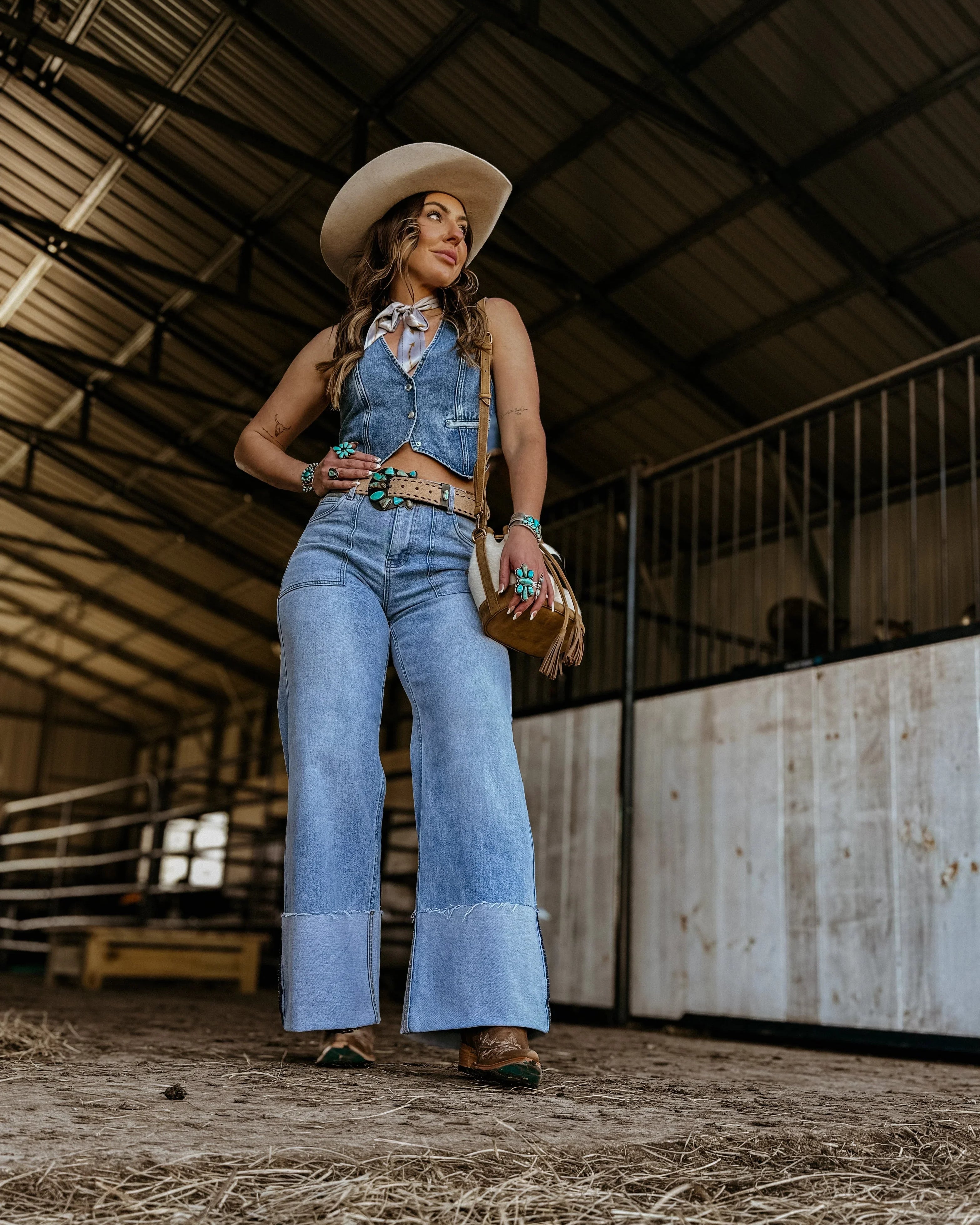 Woman in a western denim outfit with flared jeans, cowboy hat, and turquoise jewelry in a barn.