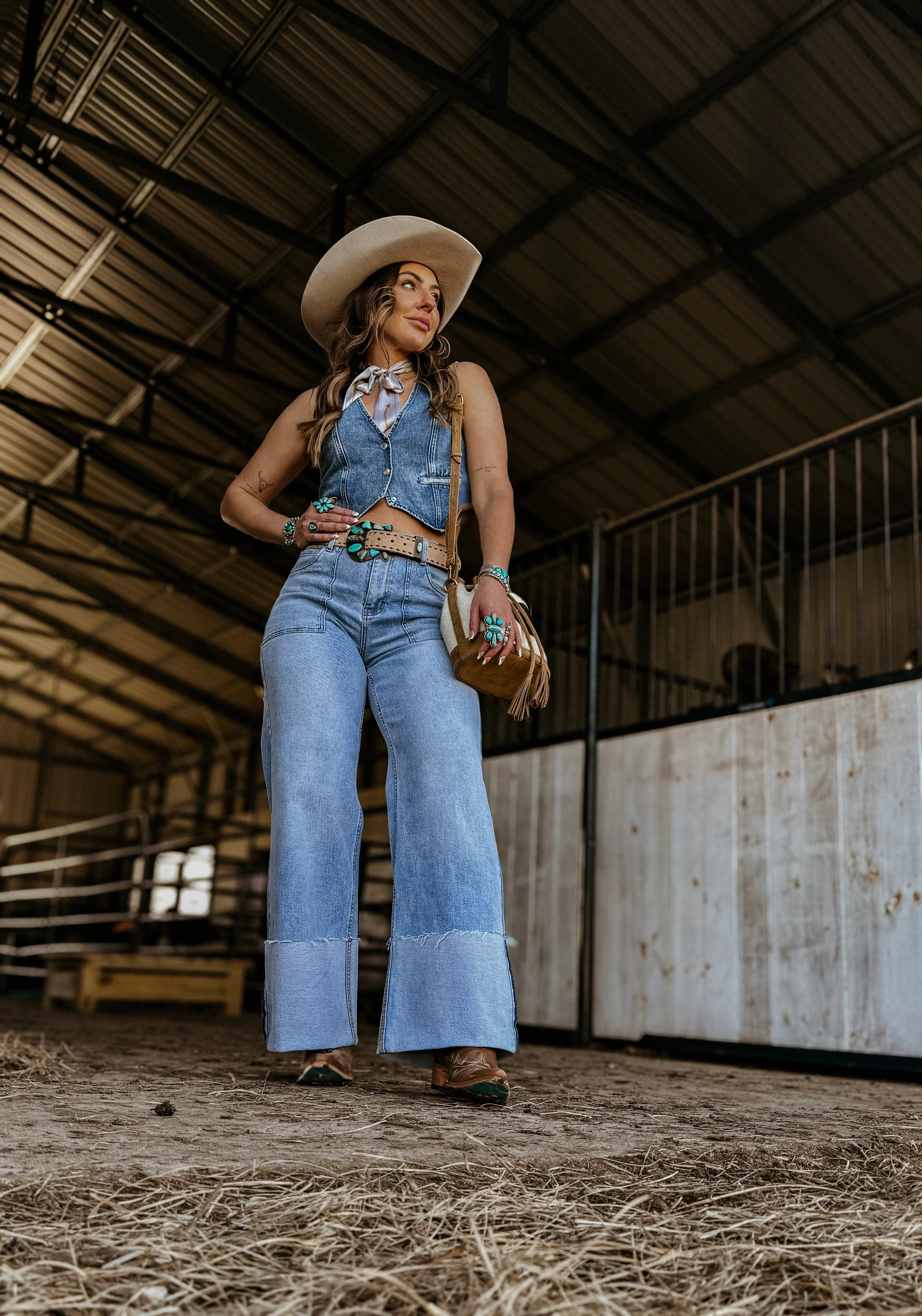 Woman in a western denim outfit with flared jeans, cowboy hat, and turquoise jewelry in a barn.