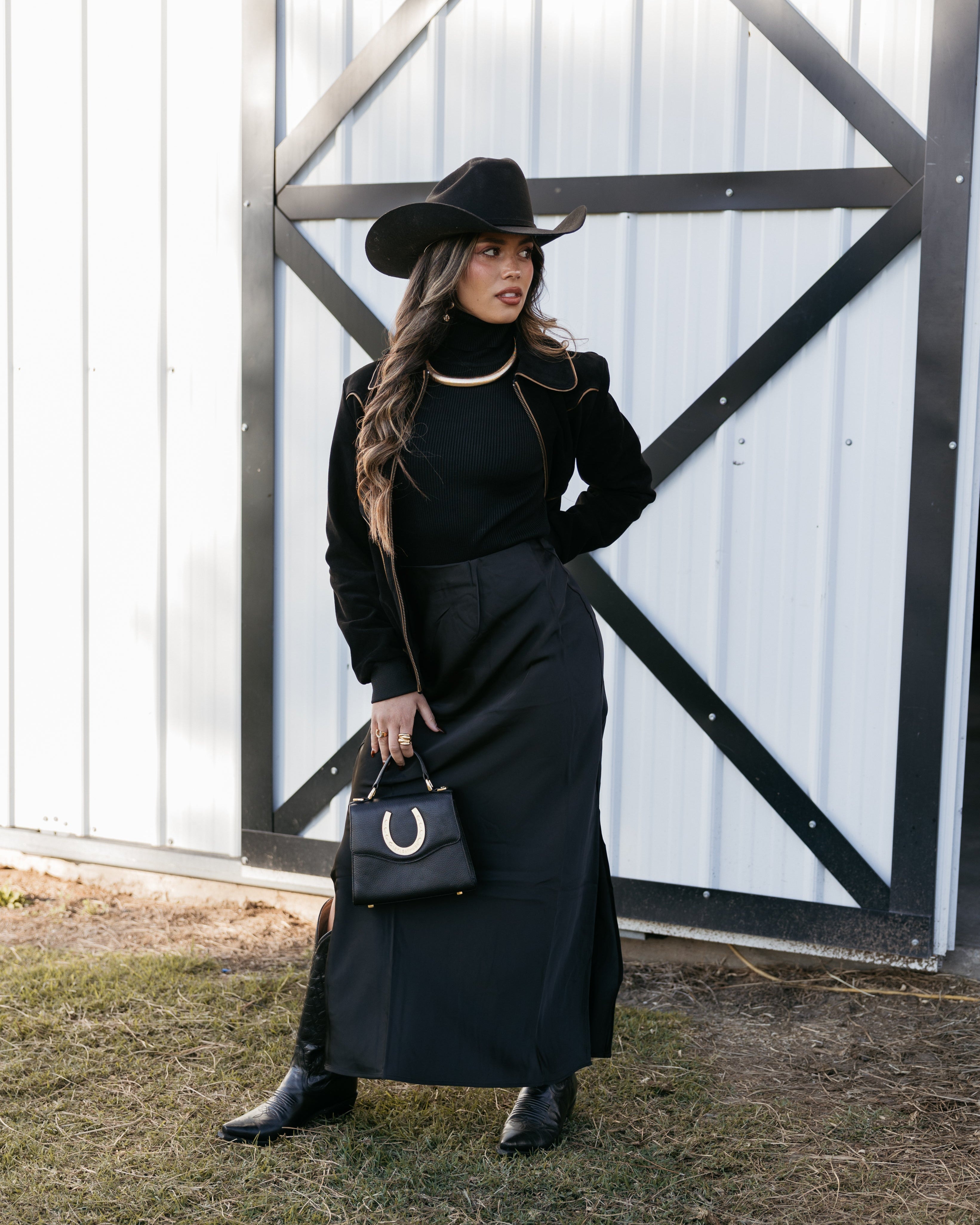 Woman in western boho outfit with black cowboy hat, long black skirt, boots, and horseshoe purse