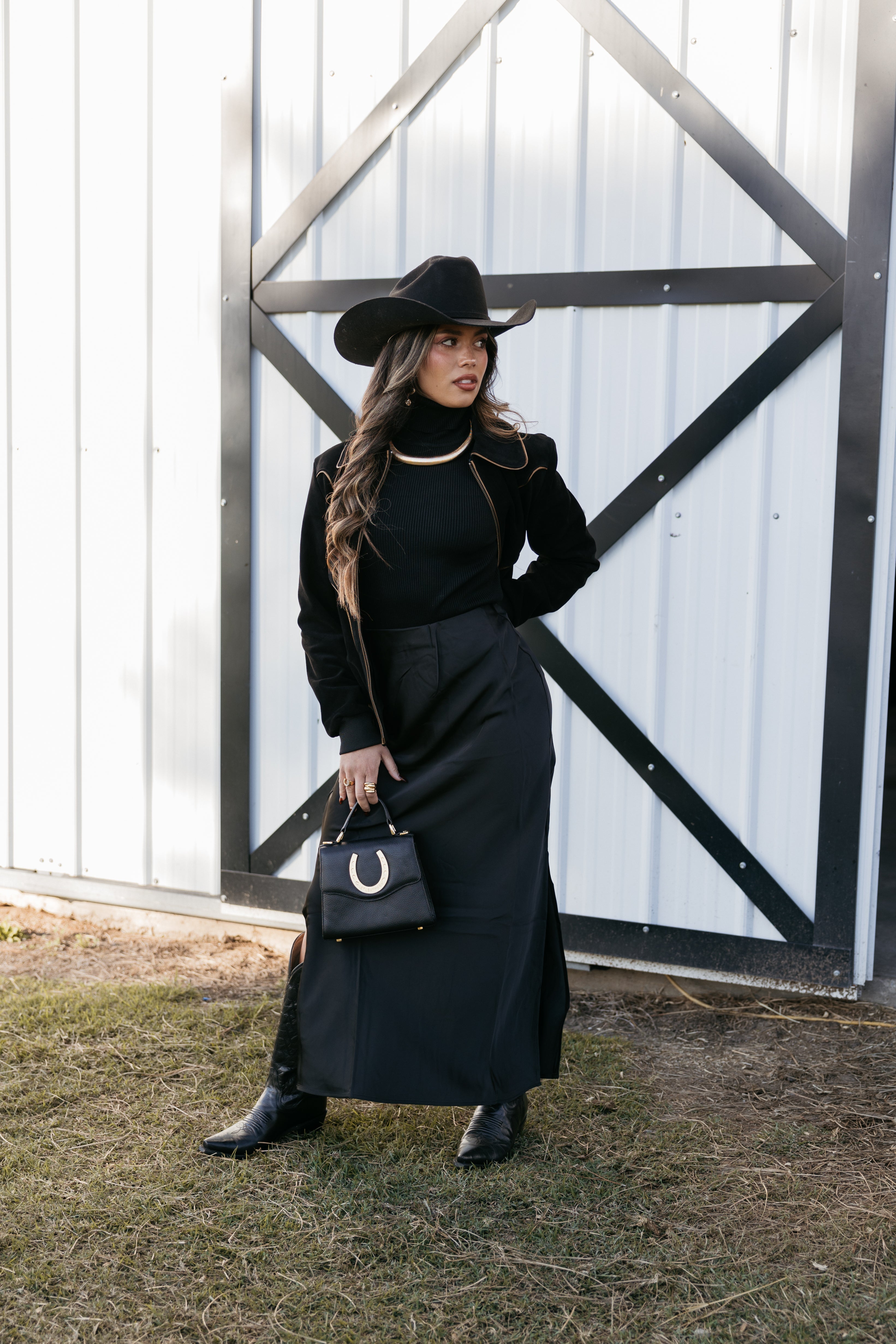 Woman in western boho outfit with black cowboy hat, long black skirt, boots, and horseshoe purse
