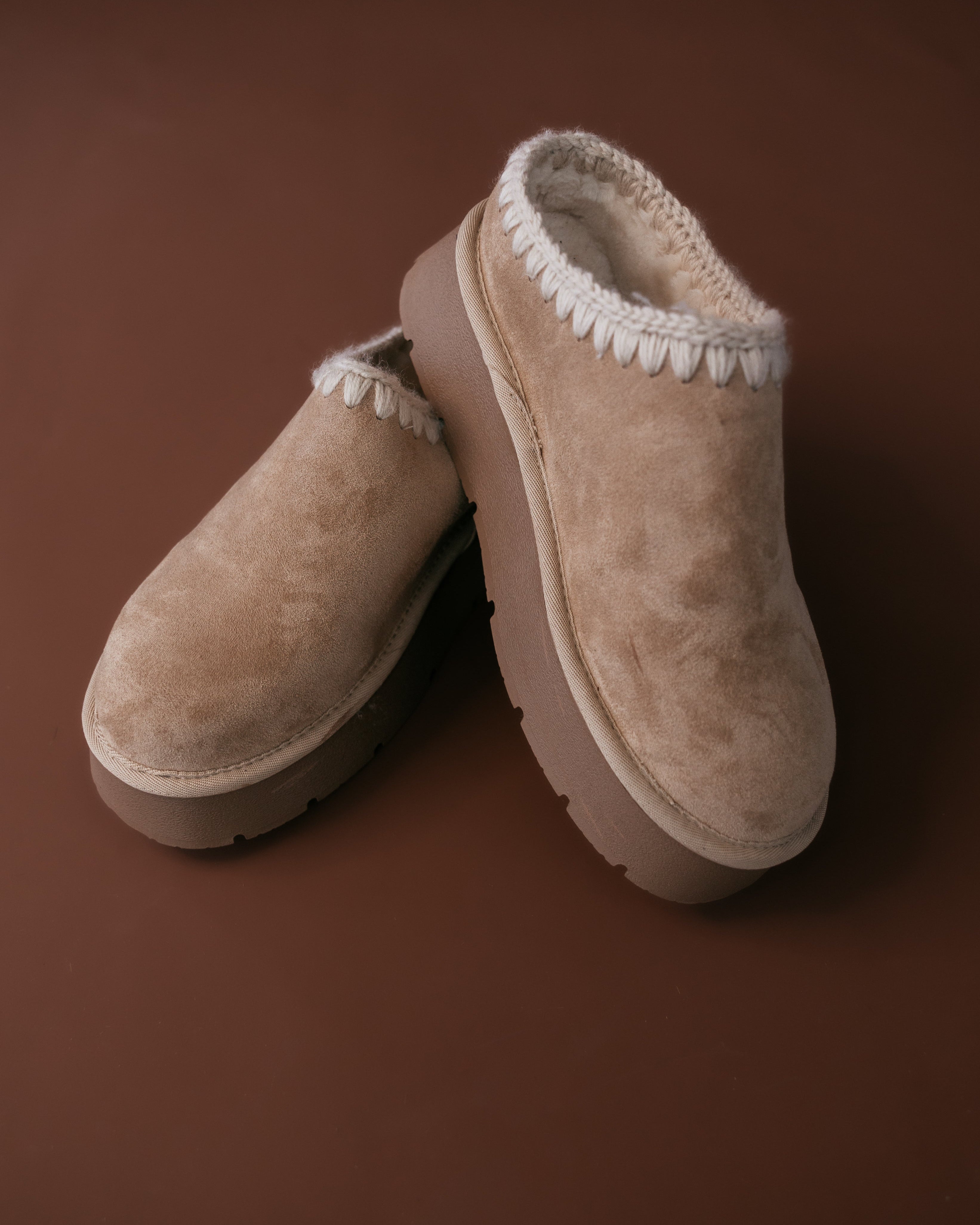 Pair of beige slippers with white fur trim on a brown background