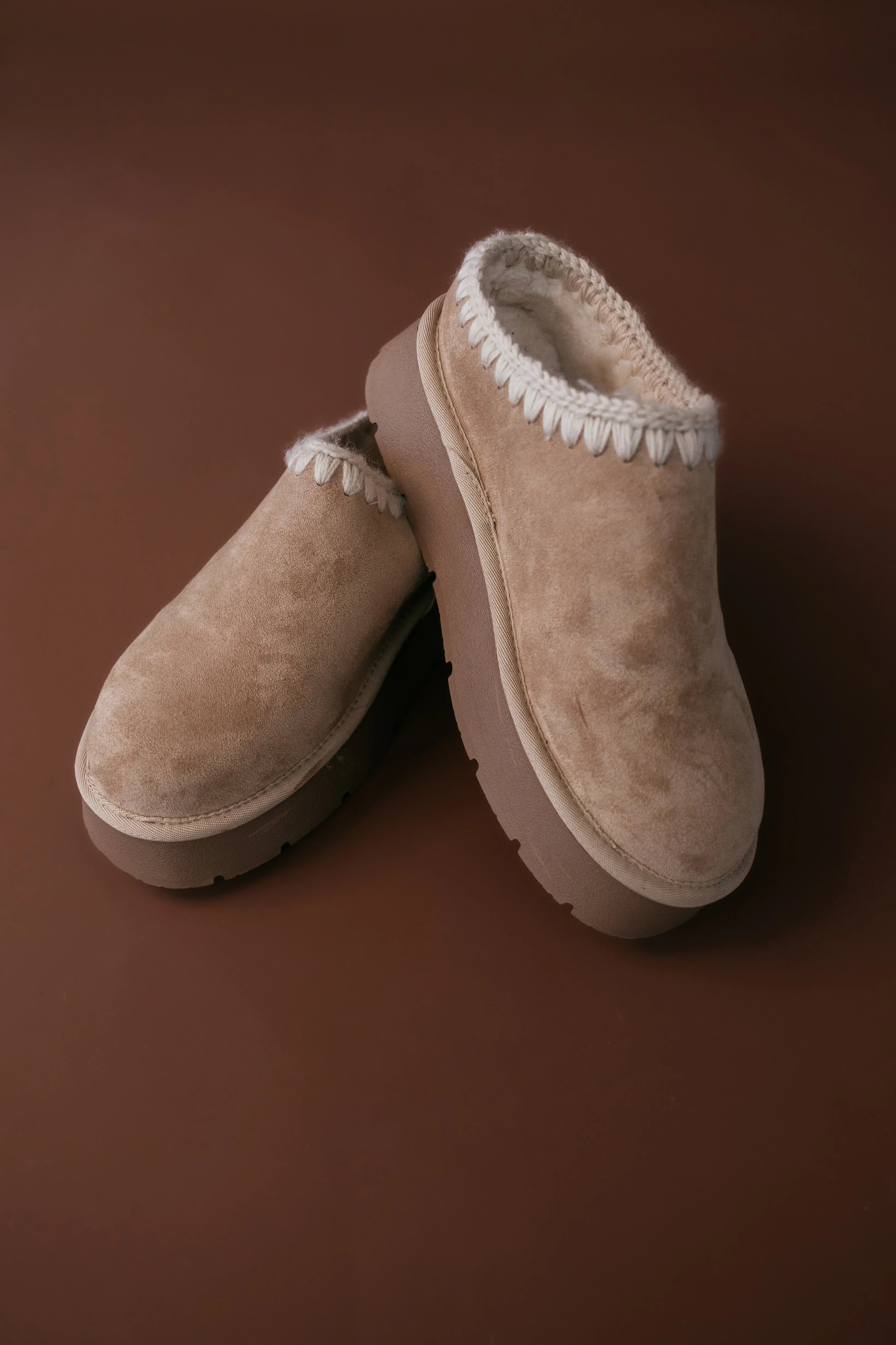 Pair of beige slippers with white fur trim on a brown background