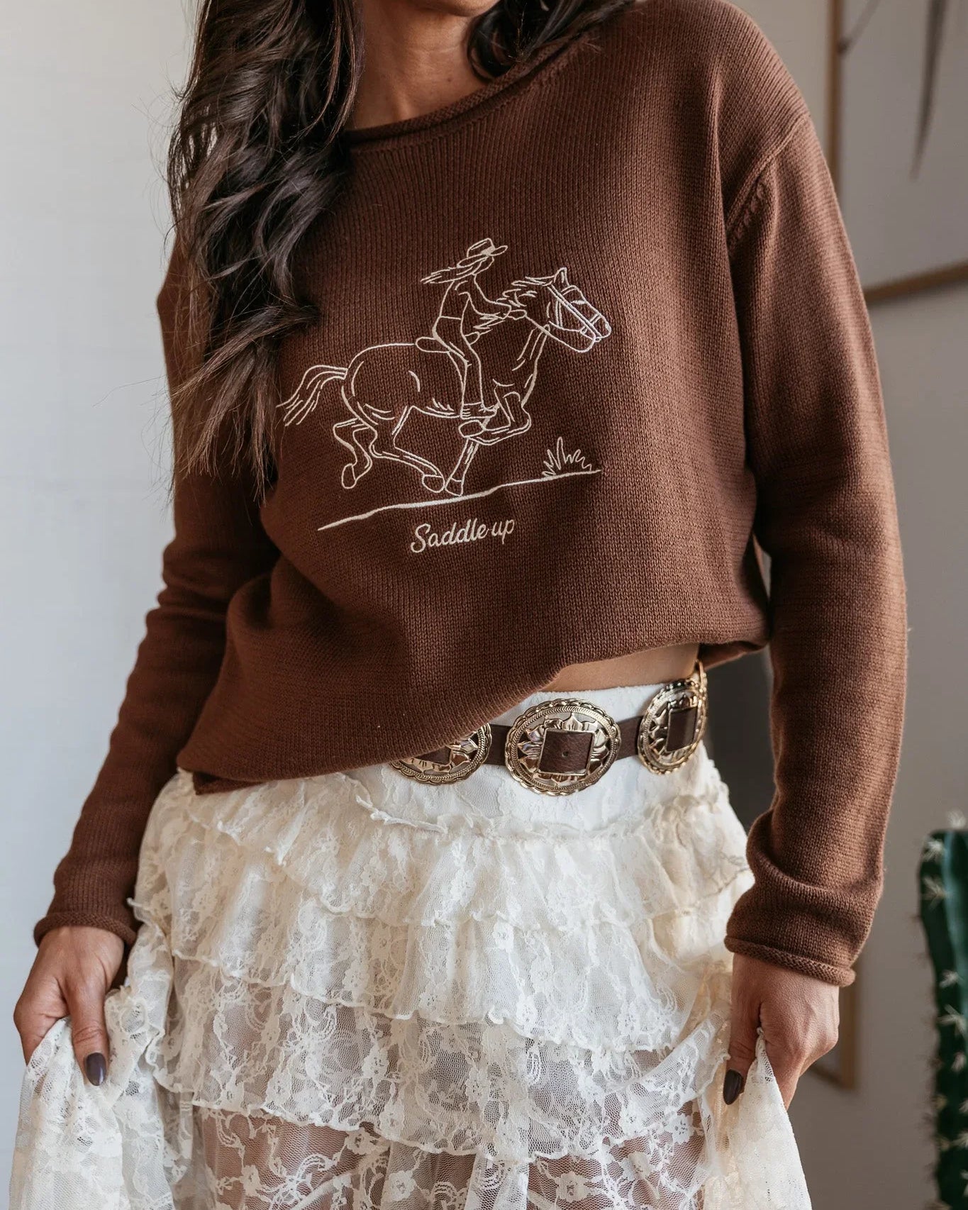 Woman in brown western sweater with horse graphic, lace skirt, and concho belt