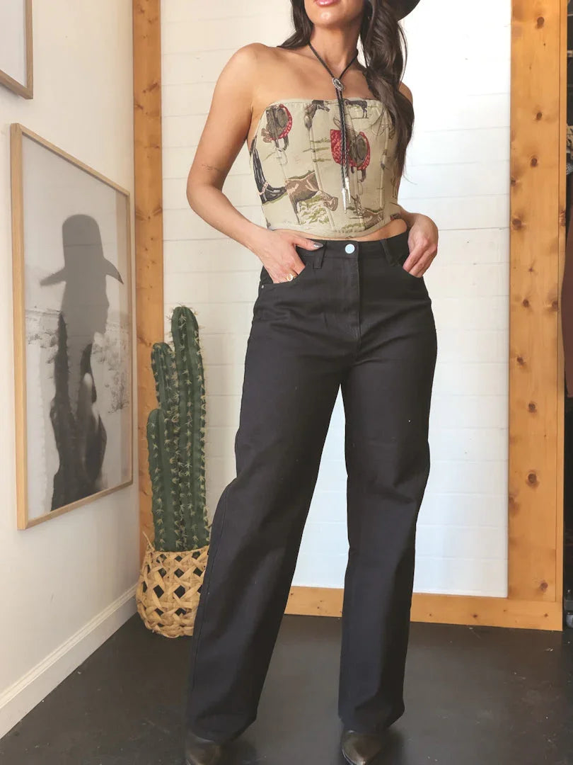 Woman in western boho outfit with rodeo print bandeau top, high-waisted black jeans, bolo tie, and cowboy hat standing indoors near cactus.